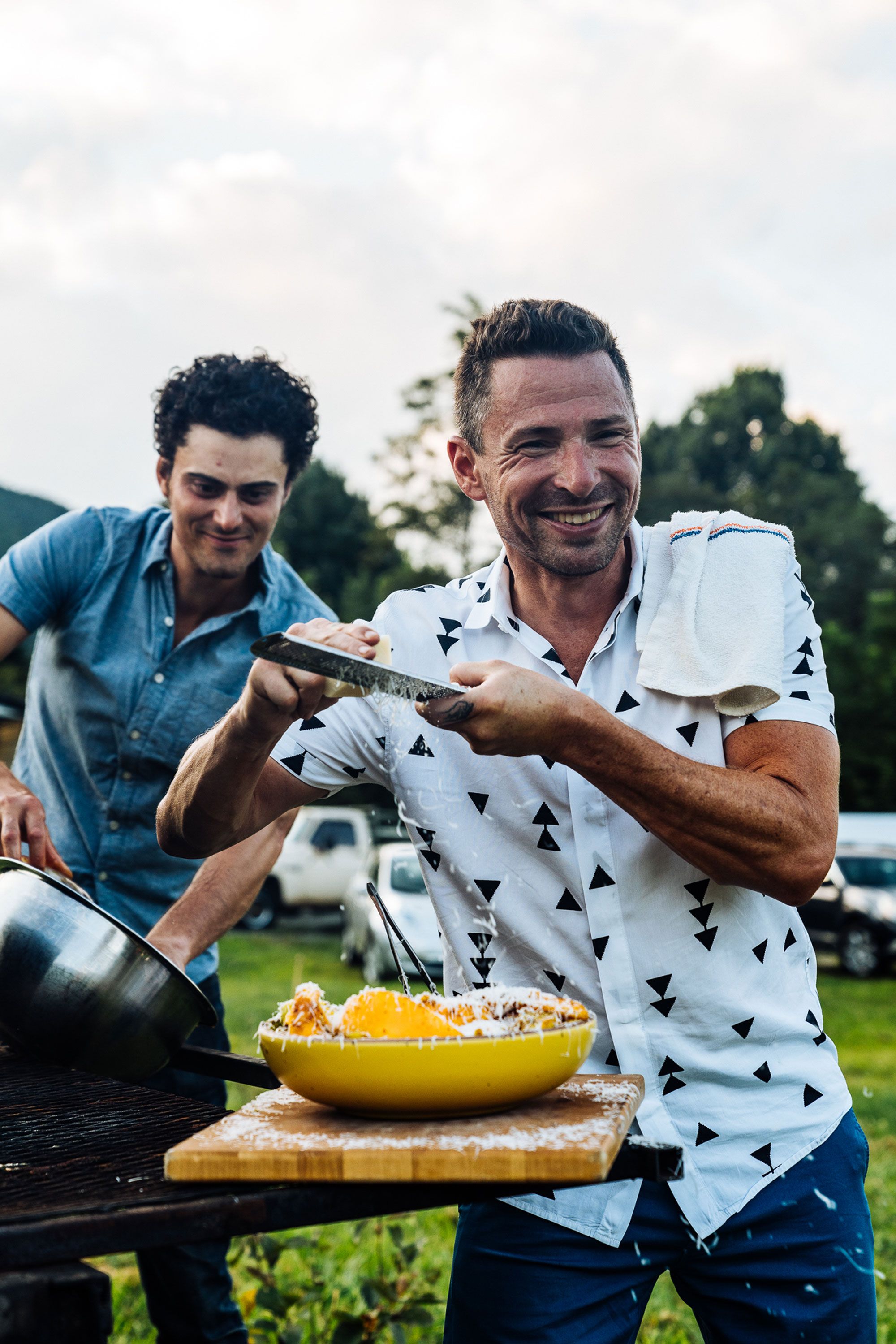 Outside, a chef is smiling and grating cheese over a large serving bowl filled with a dish