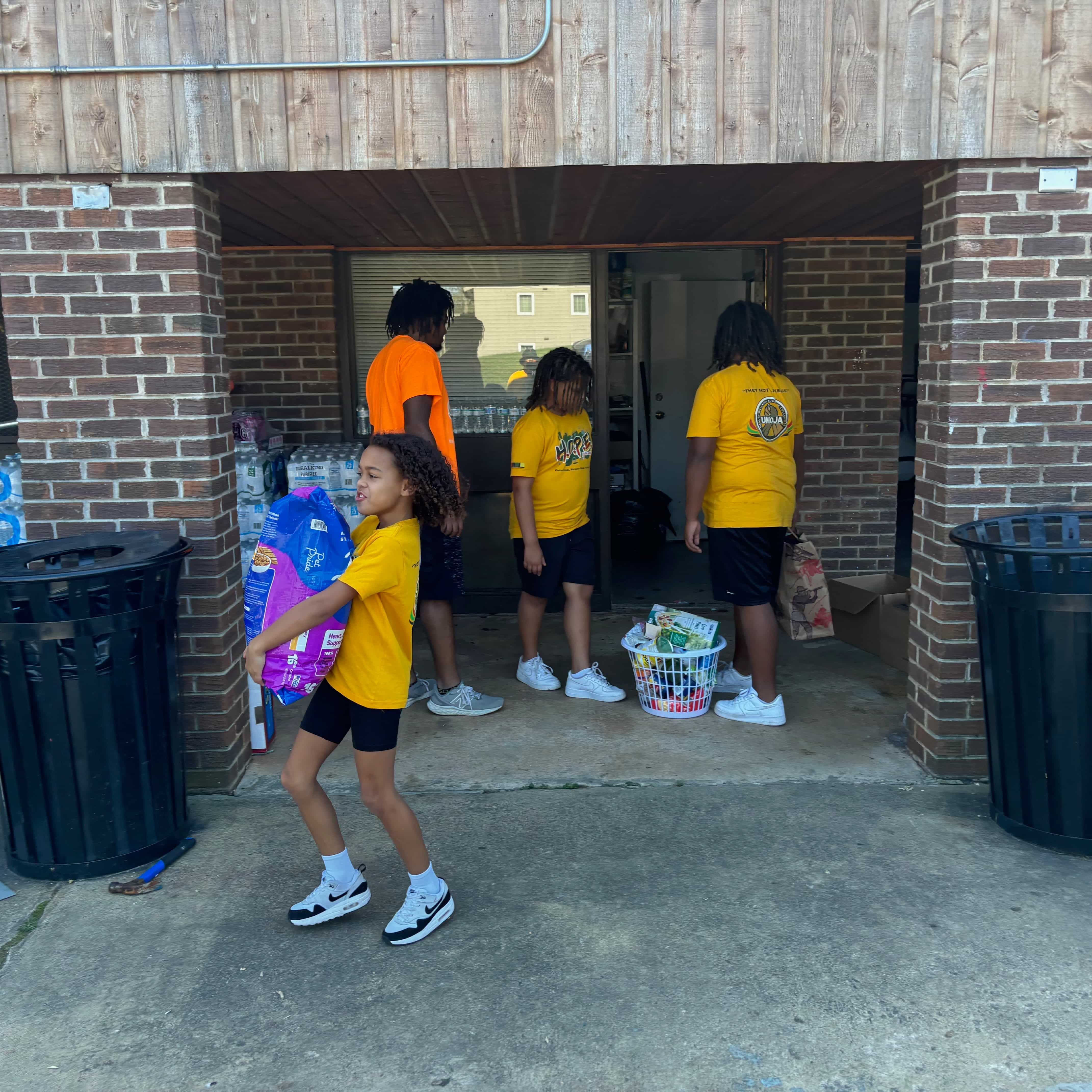 People are outside wearing yellow t-shirts and around supplies like large flats of water. A kid is holding a large bag of dog food.