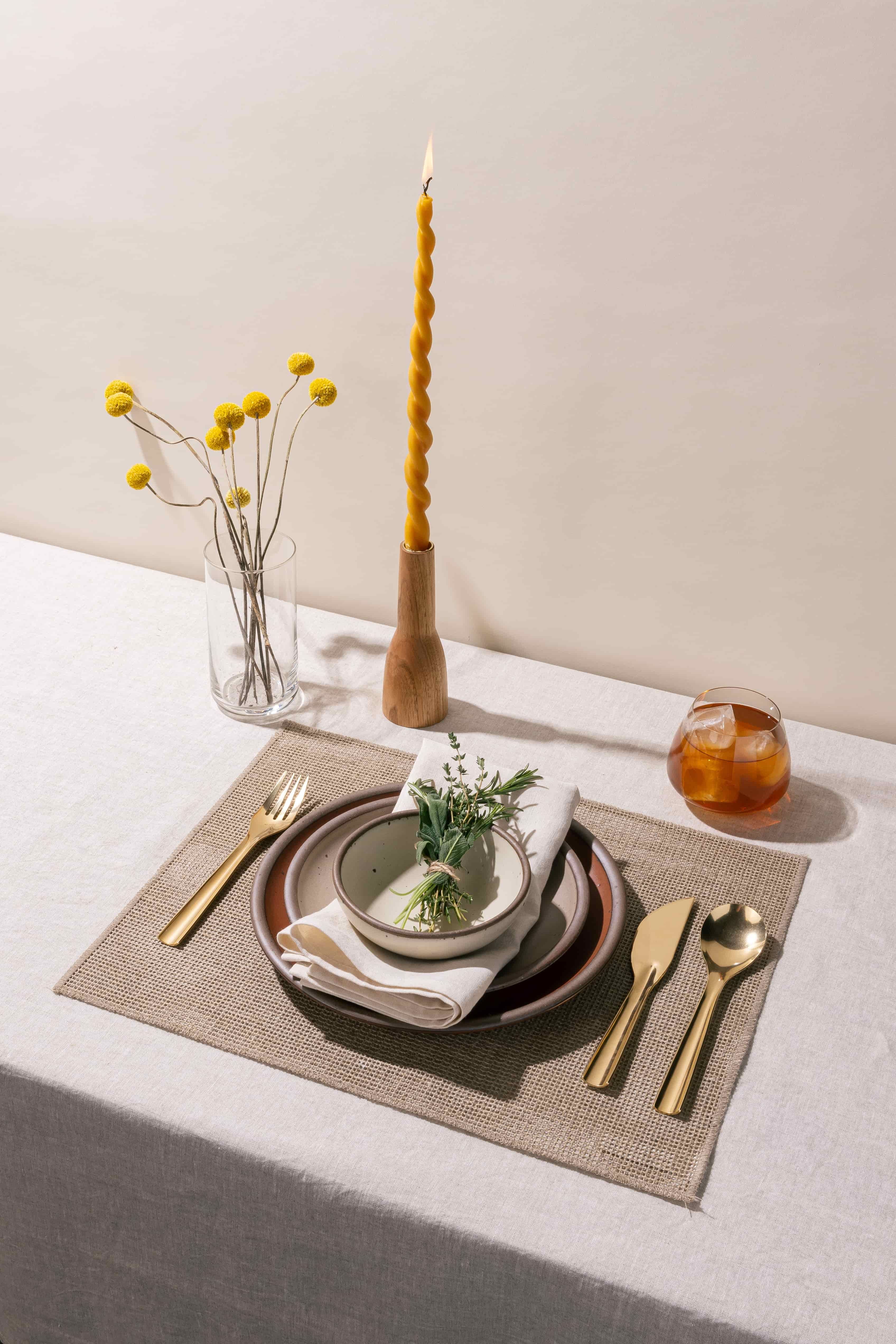 A place setting featuring ceramic plates in terracotta, neutral, and white colors, brass flatware, a burning twisted taper candle, and more.