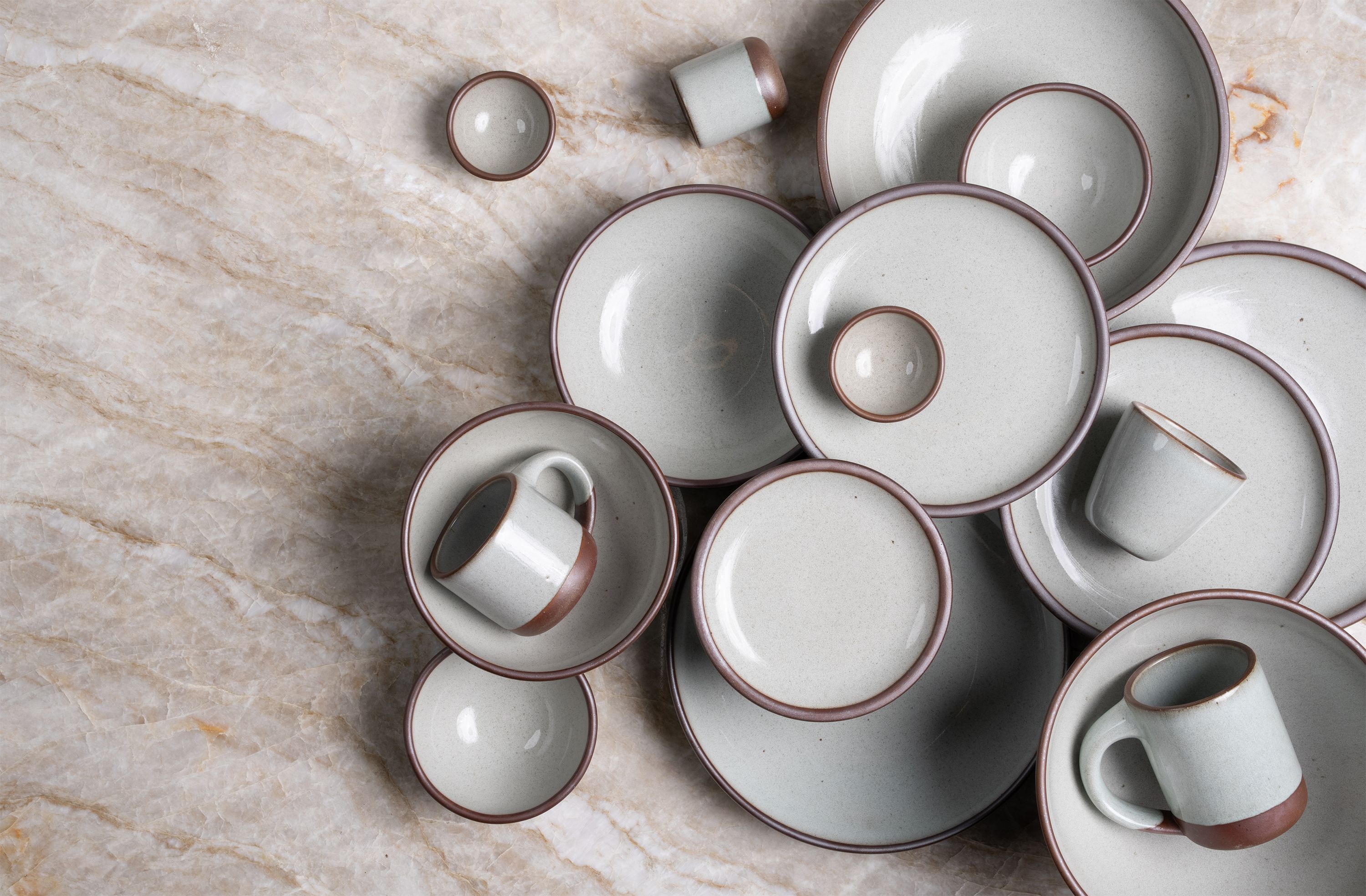 Assorted reflective icy white ceramic plates, mugs, and bowls with unglazed rims arranged on a beige marble surface.