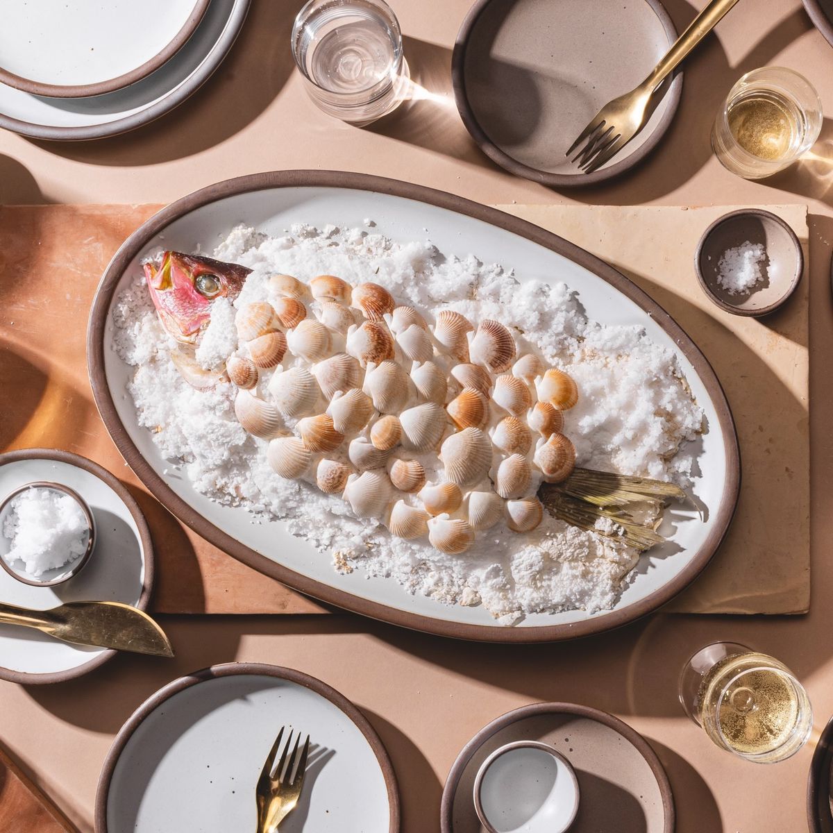 A ceramic oval platter in a cool white color with a large fish covered in salt and seashells. The platter is surrounded by plates in cool white and taupe colors and brass forks and knives.
