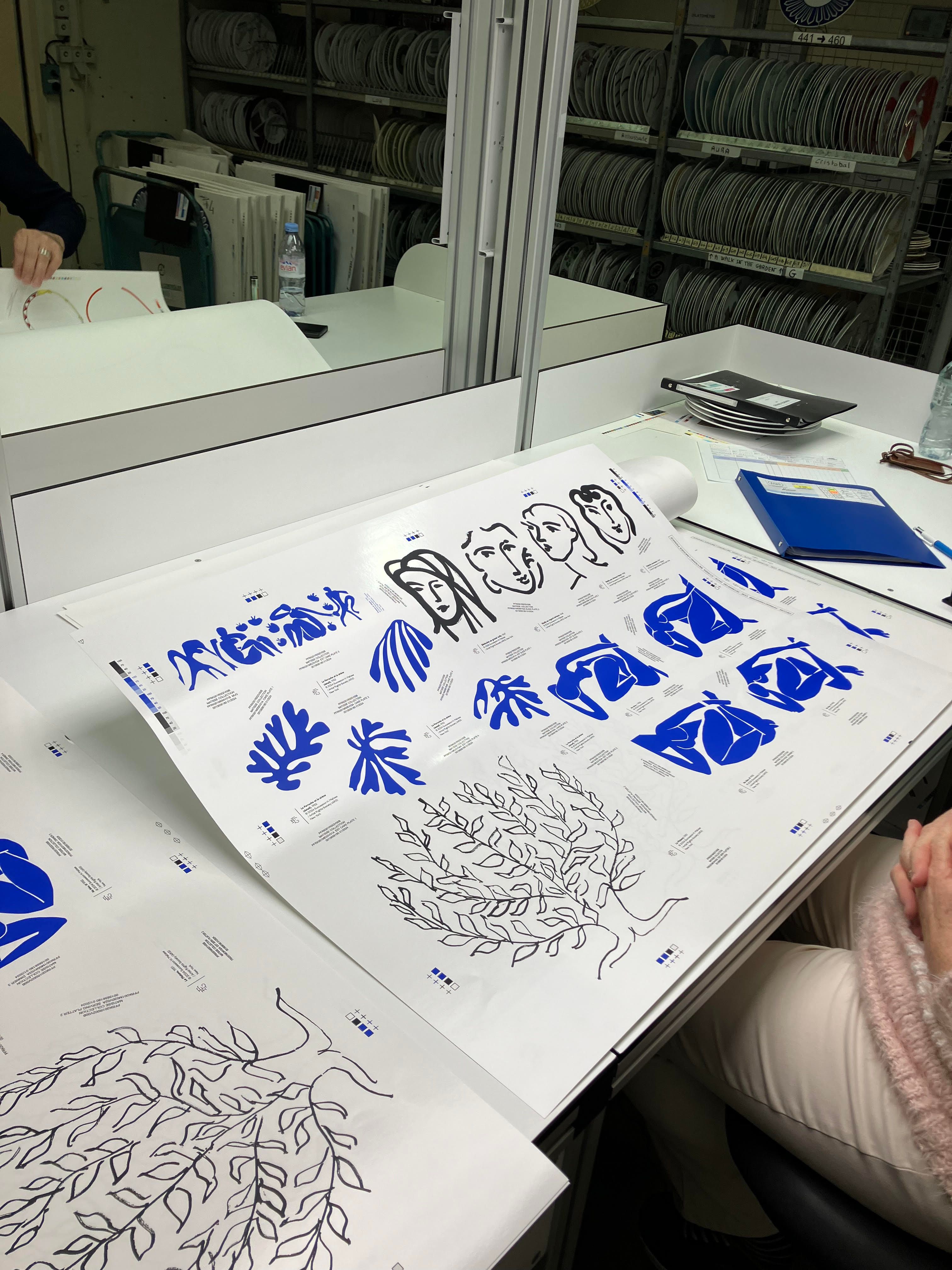Decals of Matisse's abstract paintings of blue leaves, black drawings of trees and faces, and blue cut-out art of women, in a factory on a desk.