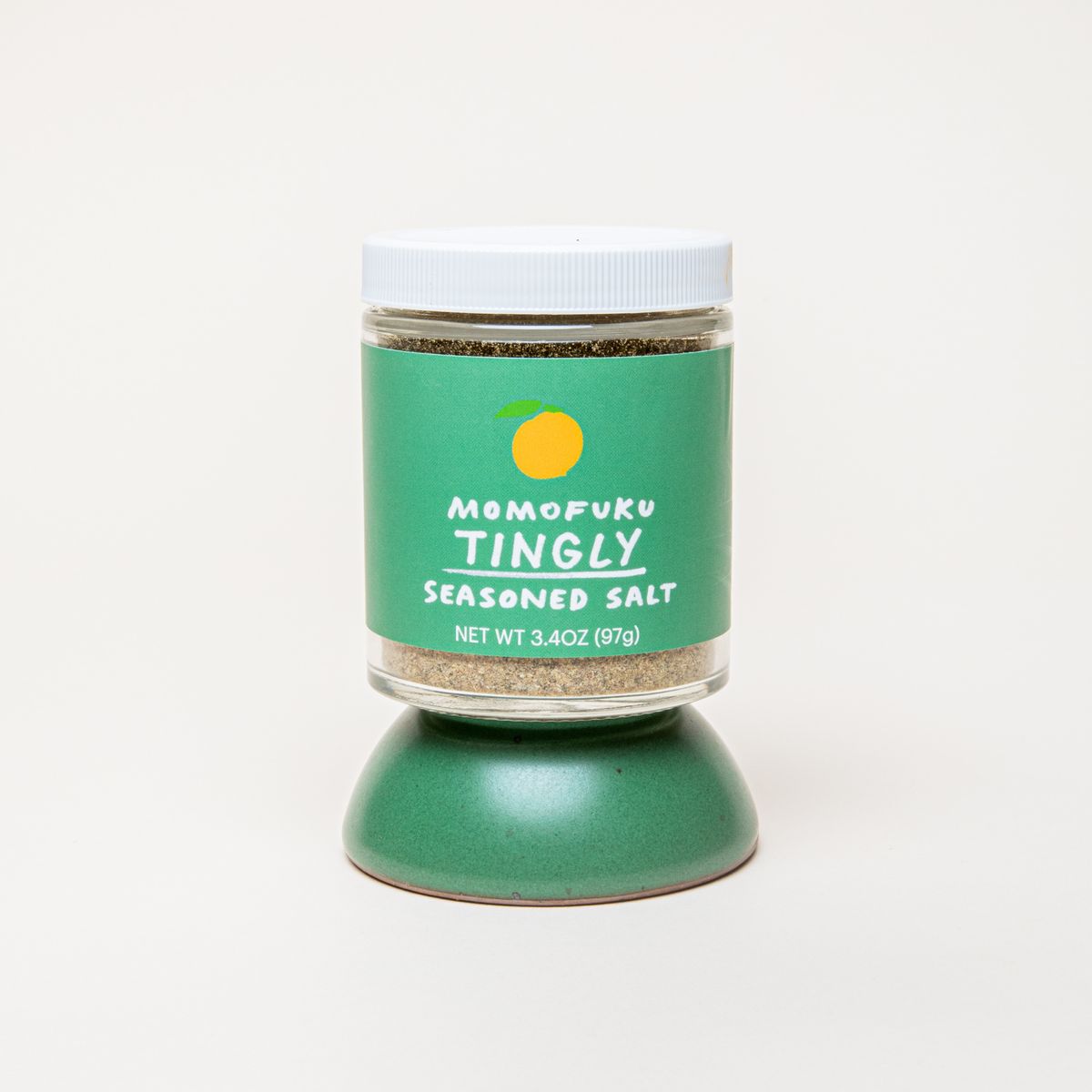 Short jar of salt with a white top and a green label that reads "Momofuku Tingly Seasoned Salt", sitting on an upside down small ceramic bowl