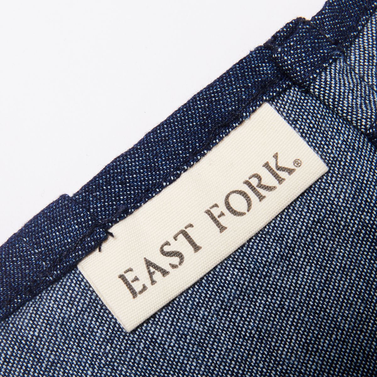 White tag sewn into the inside of a denim blue apron that reads 'East Fork'