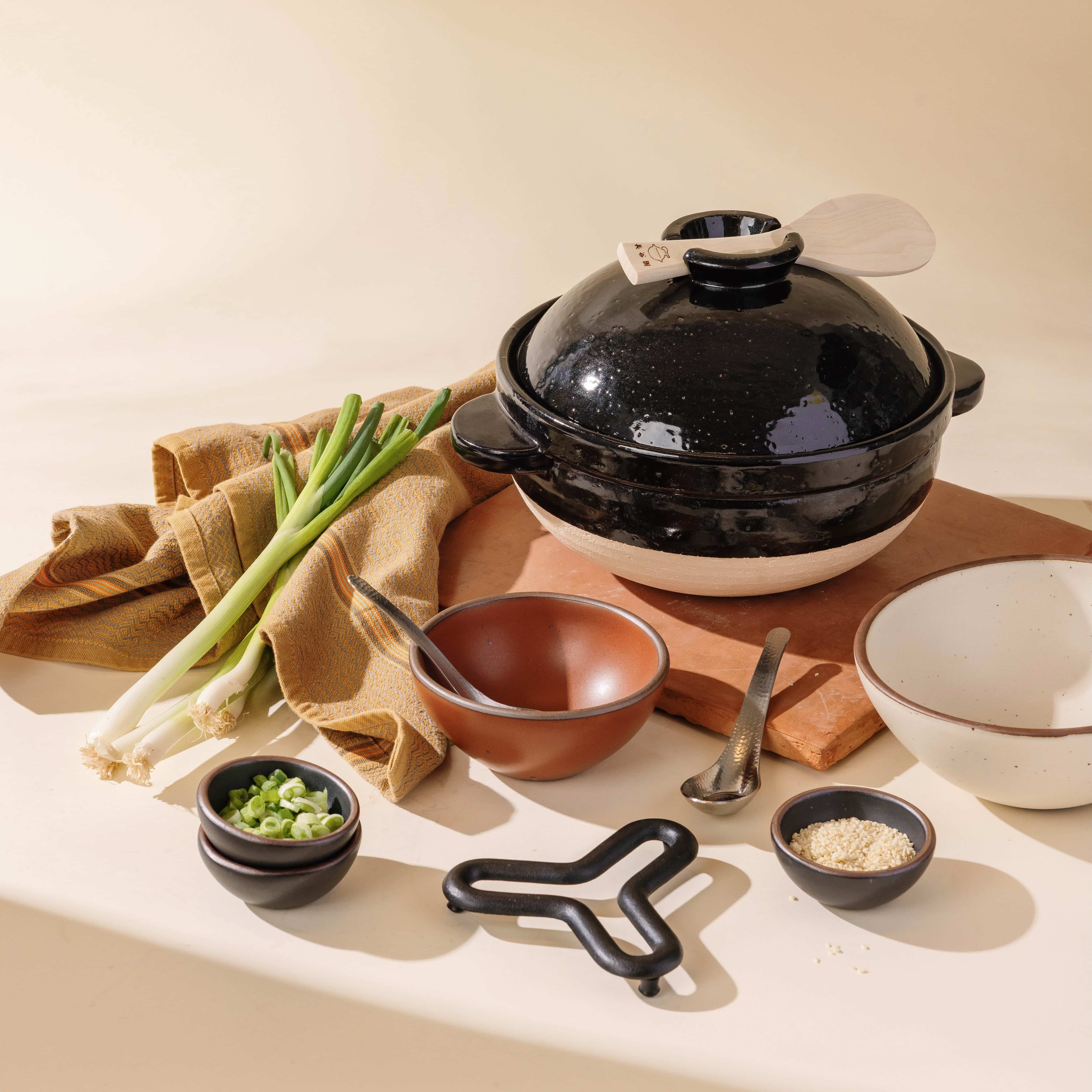 Black Donabe styled with fresh scallions, sesame, and serving pieces for a modern, elevated kitchen vignette.