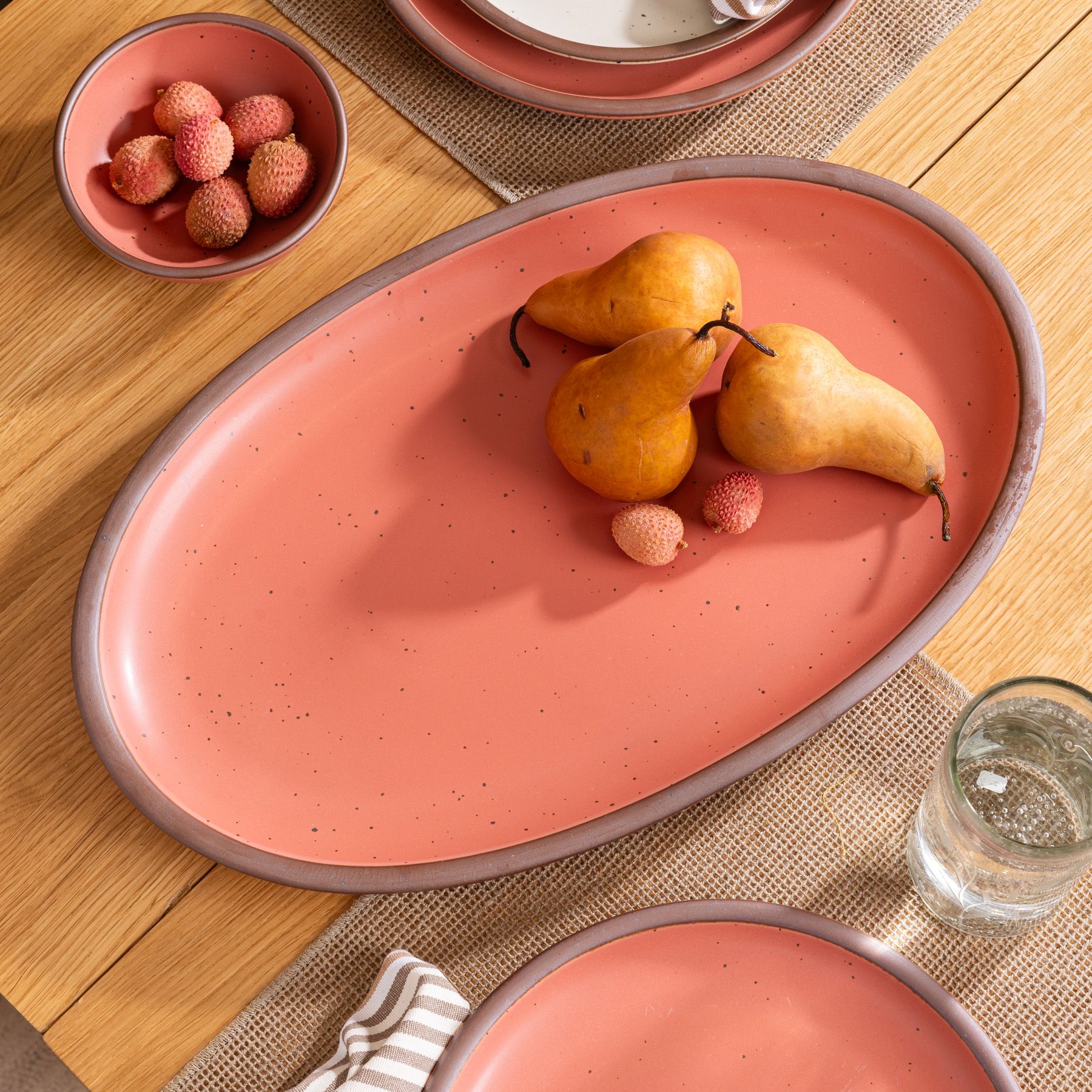 Watermelon pink oval serving platter with pears and lychees on a wood table set with matching dishes and a glass of water.
