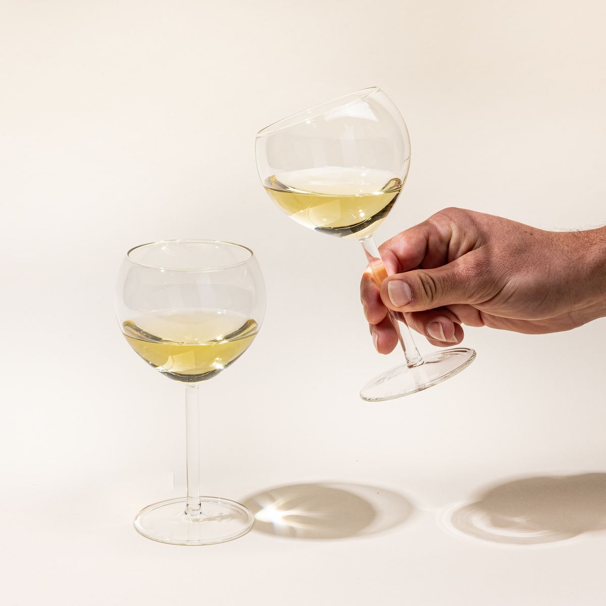 Two simple round clear wine glasses filled with white wine. A hand holds one glass up.