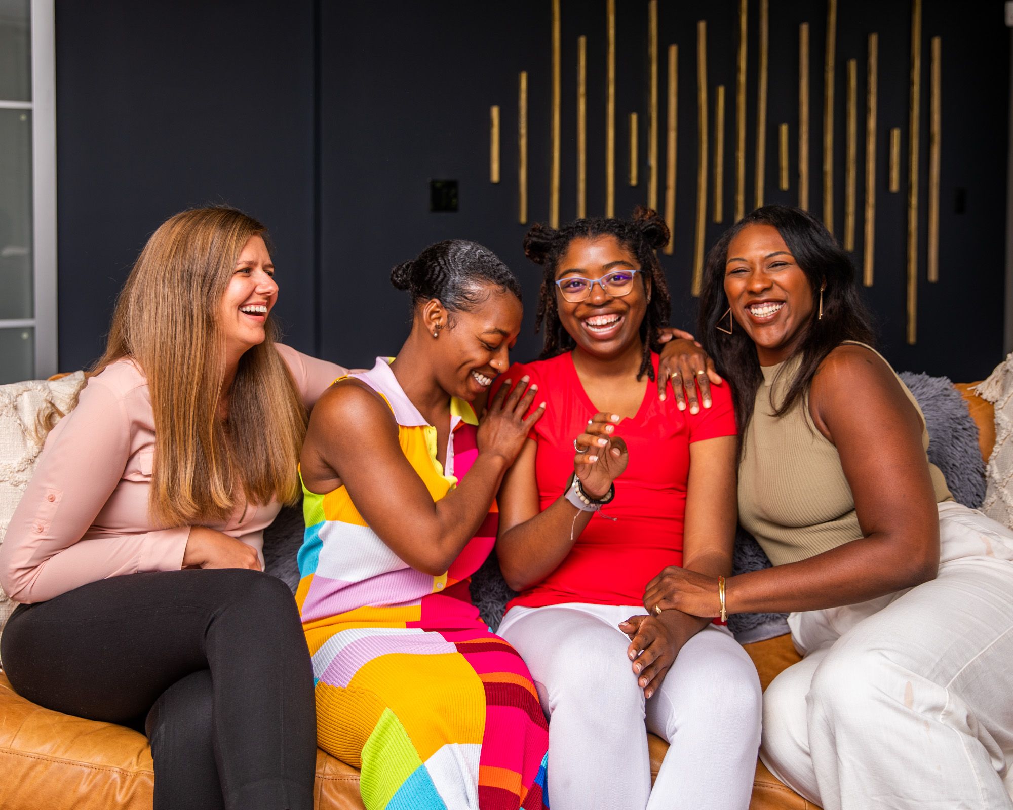 Four women sitting on a couch close together hugging and smiling with each other against a black and gold background.