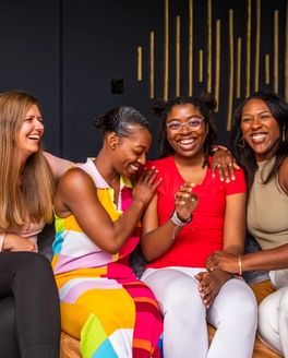 Four women sitting on a couch close together hugging and smiling with each other against a black and gold background.