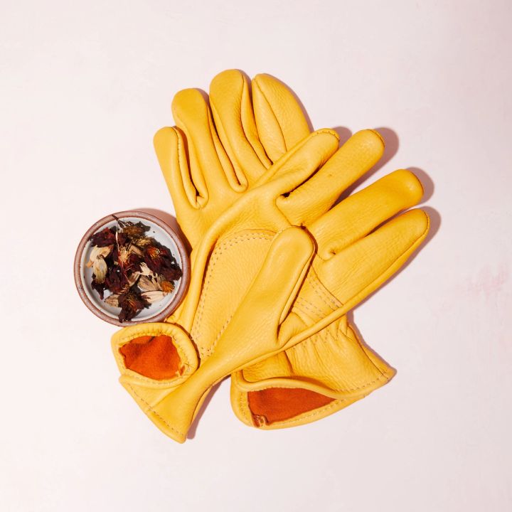 2 Buttery yellow deerskin garden gloves with brown orange interior laying flat, with a small bowl of flowers