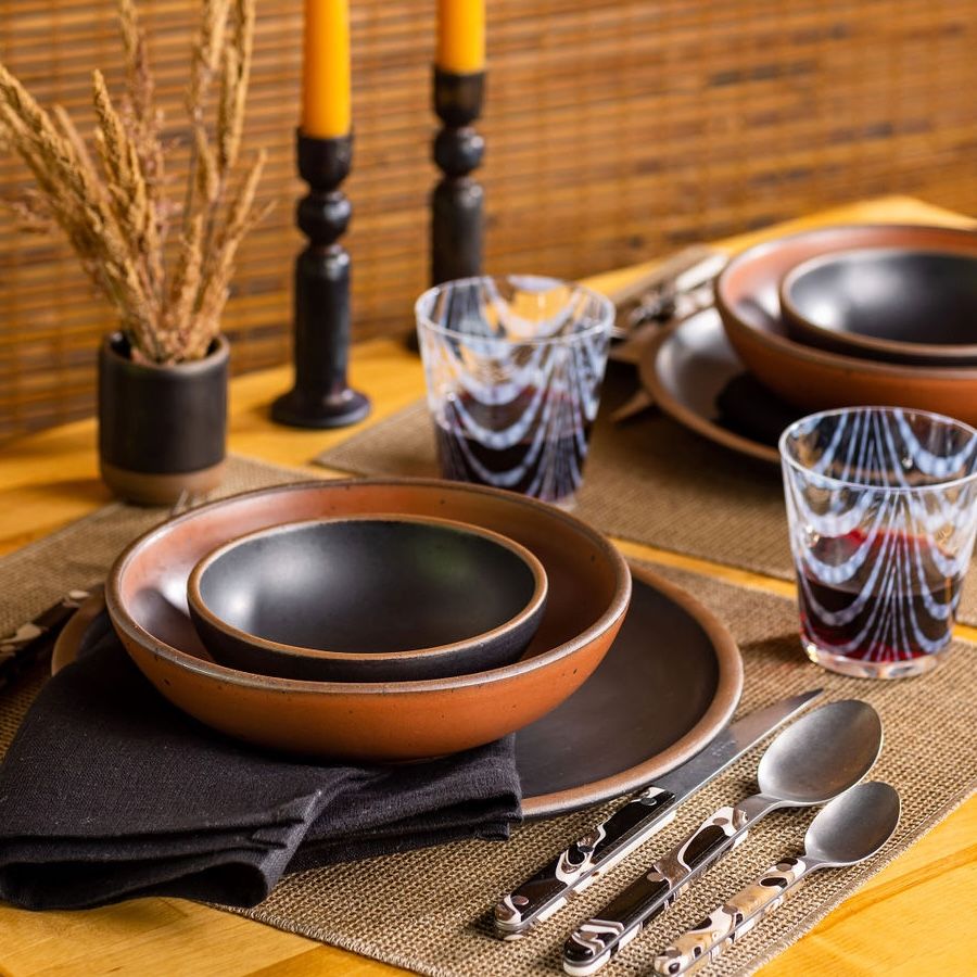 An elegant place setting featuring ceramic dinnerware in black and terracotta colors, marble handled flatware, black linen napkin, and artful marbled tumbler glasses.