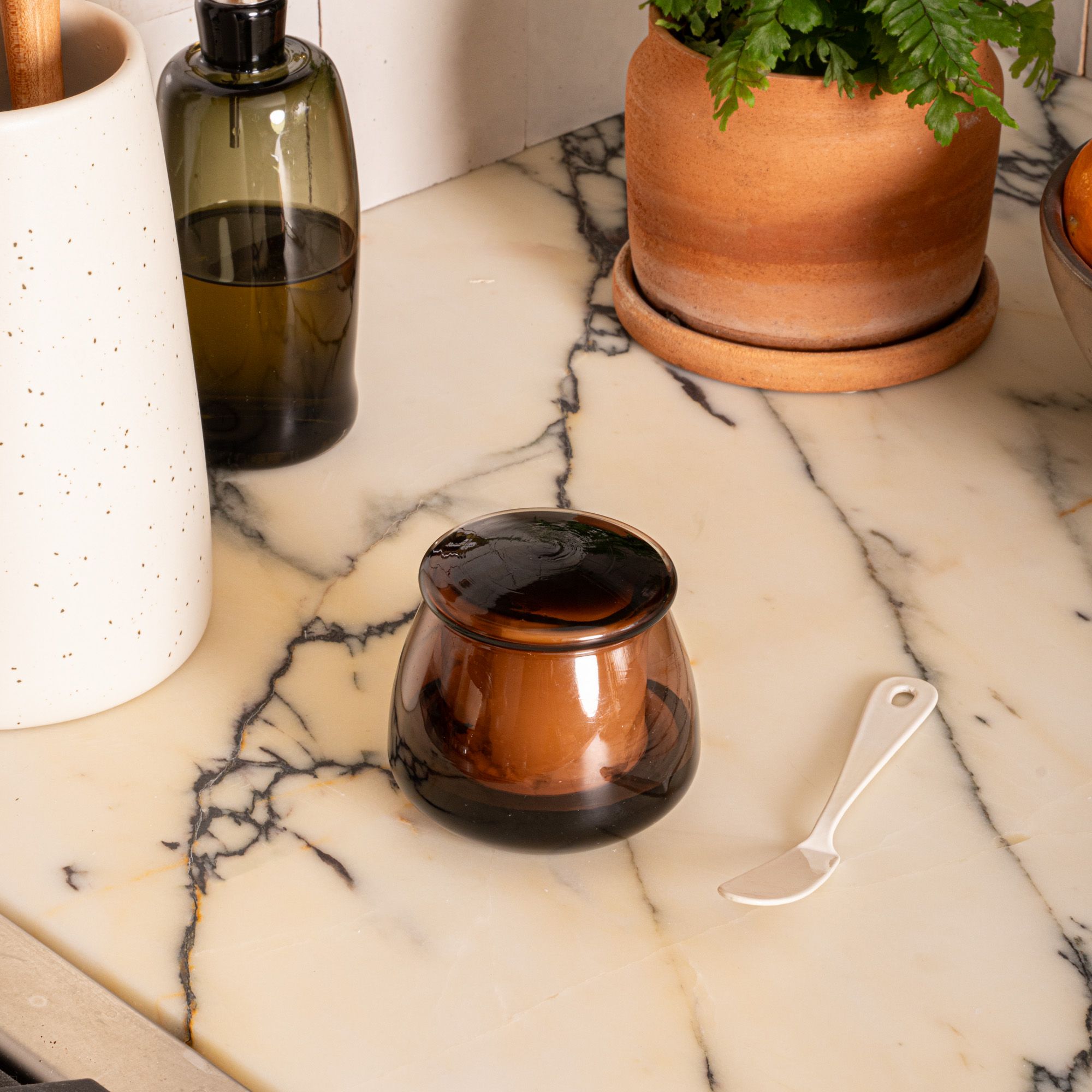 On a kitchen countertop is a brown glass butter keeper filled with butter, next to a butter spreader, potted plant, cruet, and vase.