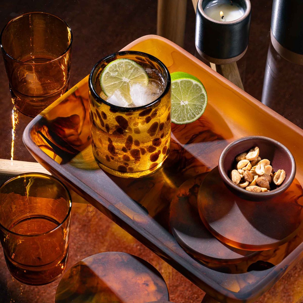 A rectangular tortoiseshell tray with curved corners filled with tortoiseshell coasters, and a tortoiseshell tumbler with a cocktail.