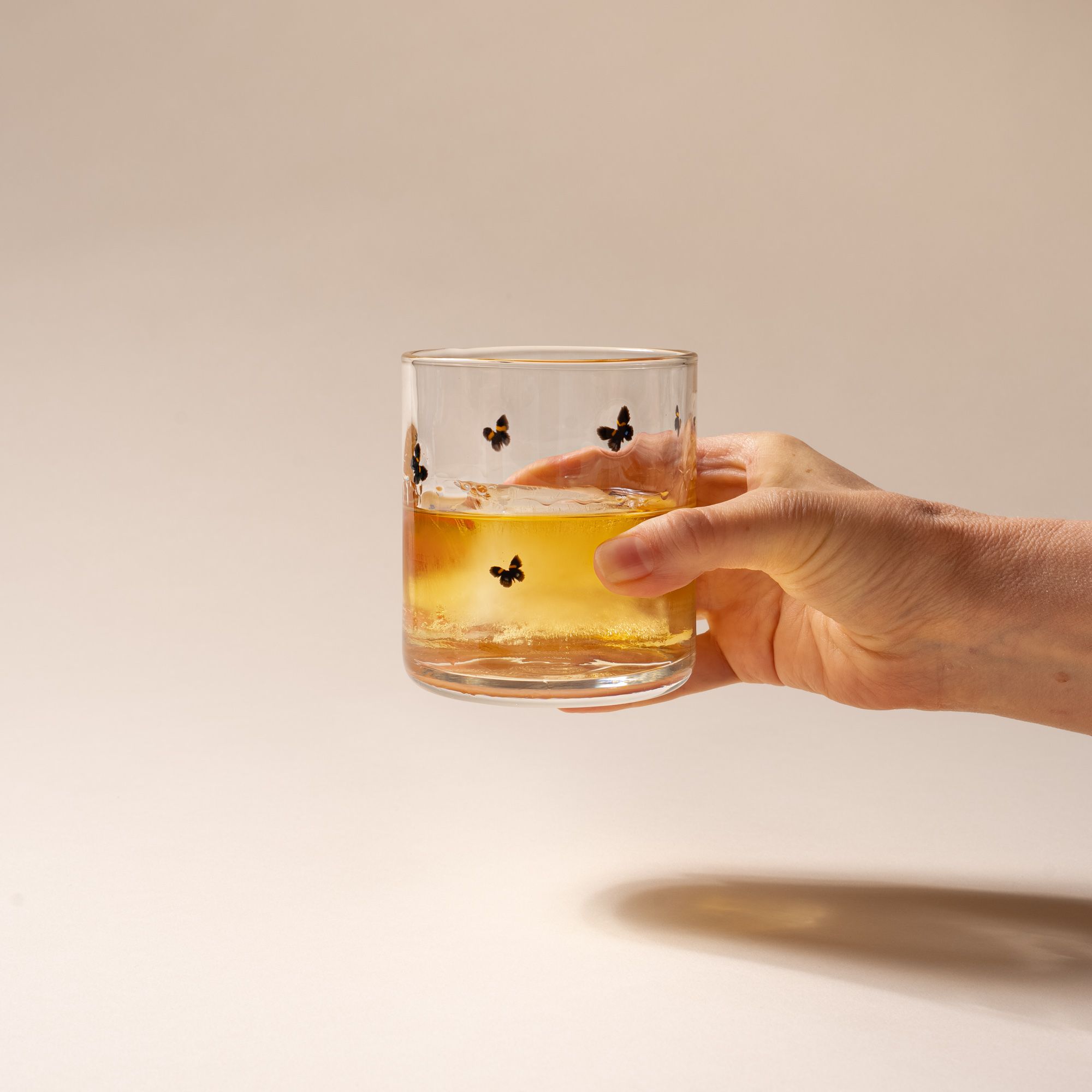 A hand holds a clear glass with a ring of butterflies on a cream background, with a drink inside.