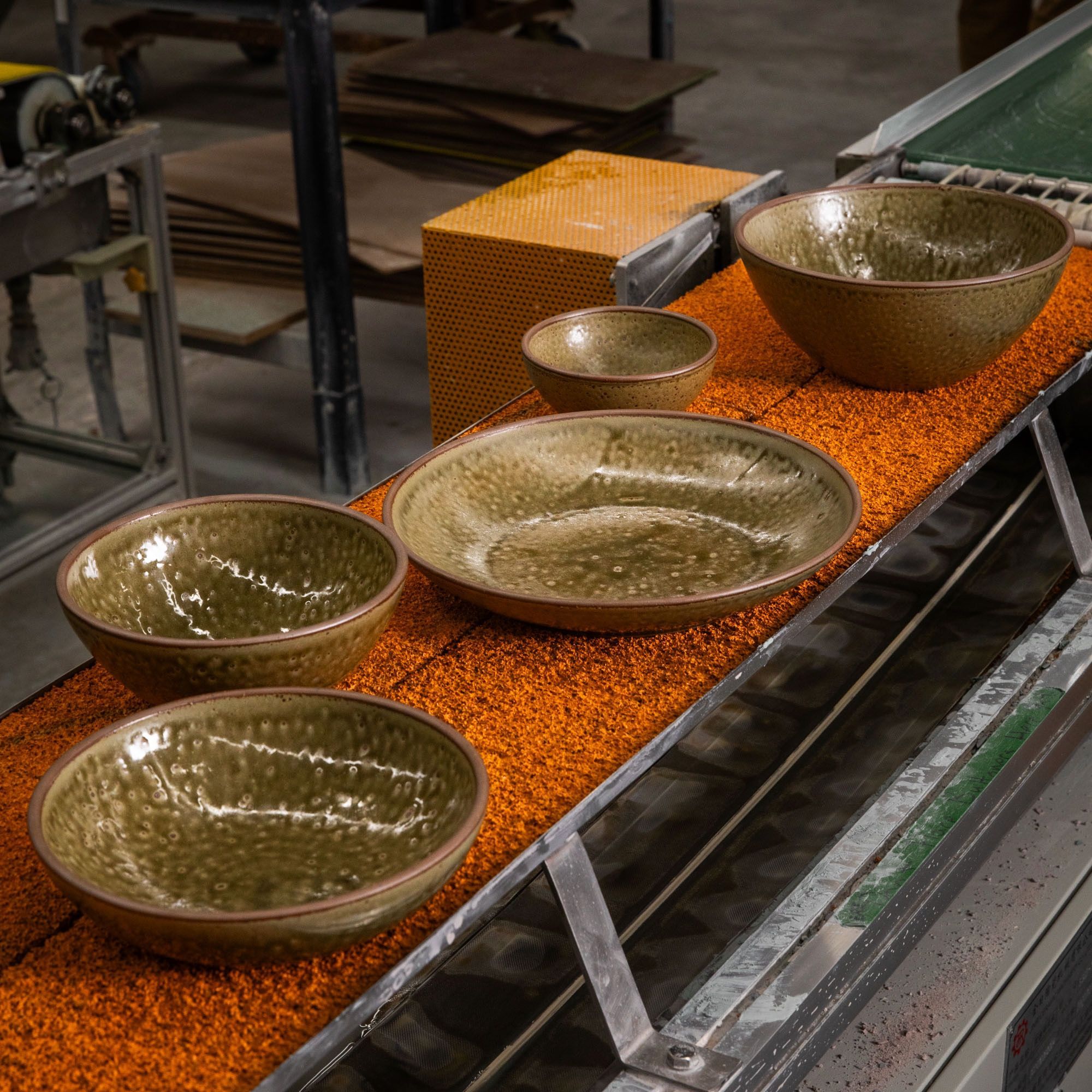 In a warehouse setting, five ceramic bowls of various sizes in a mossy earthy green color are on an orange assembly line.