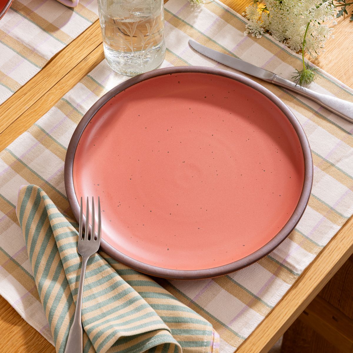 Watermelon pink ceramic dinner plate with striped napkin and silverware on a plaid runner, next to a glass of water and white flowers.