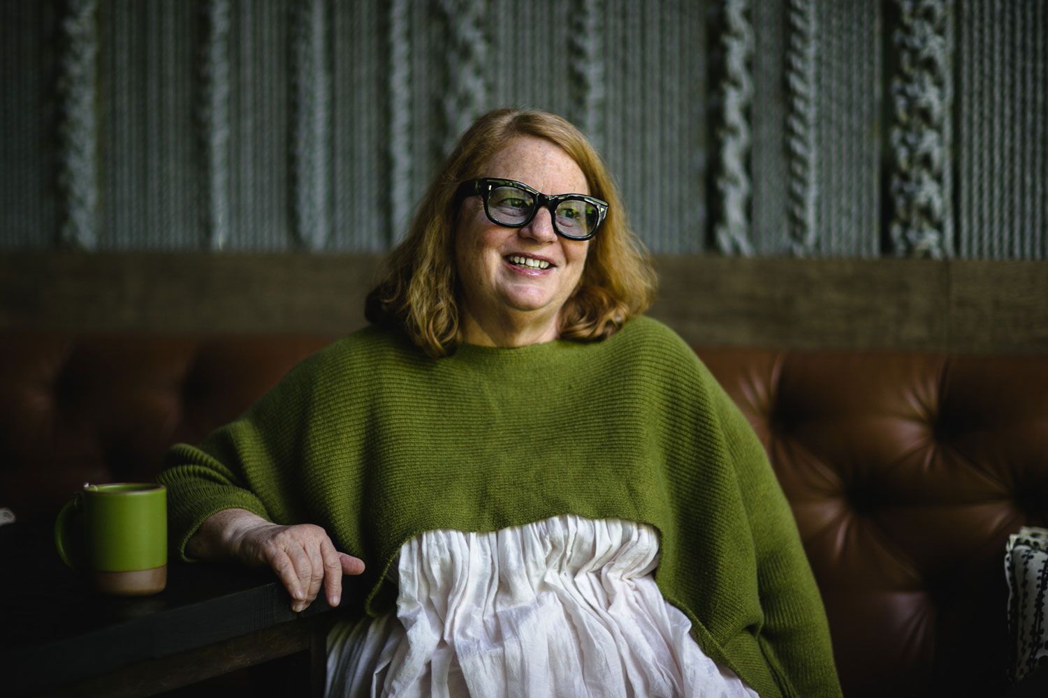 Anne Quatrano sits at a table and is smiling wearing glasses and a fern green shirt