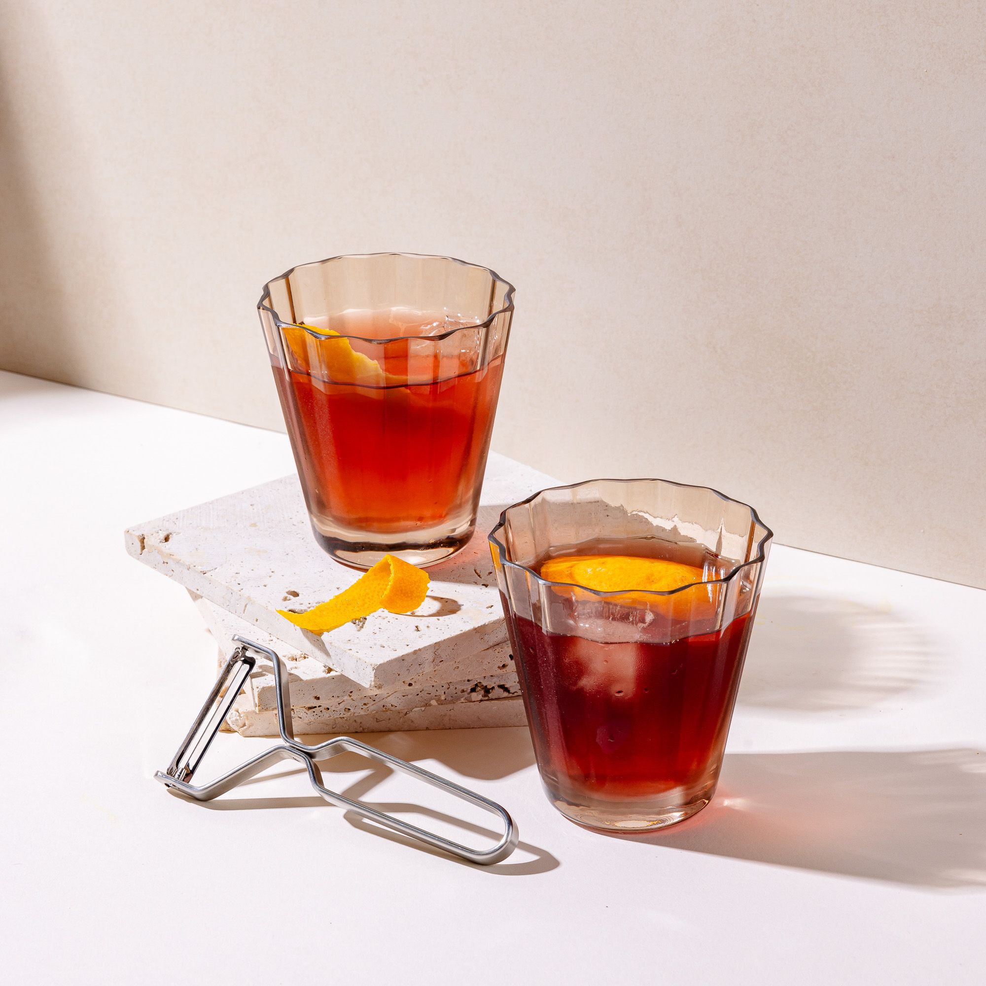 Styled two transparent light amber glasses with wide grooves on the side, filled with a cocktail and orange slice. Next to the glasses are a orange slice and stainless steel peeler.