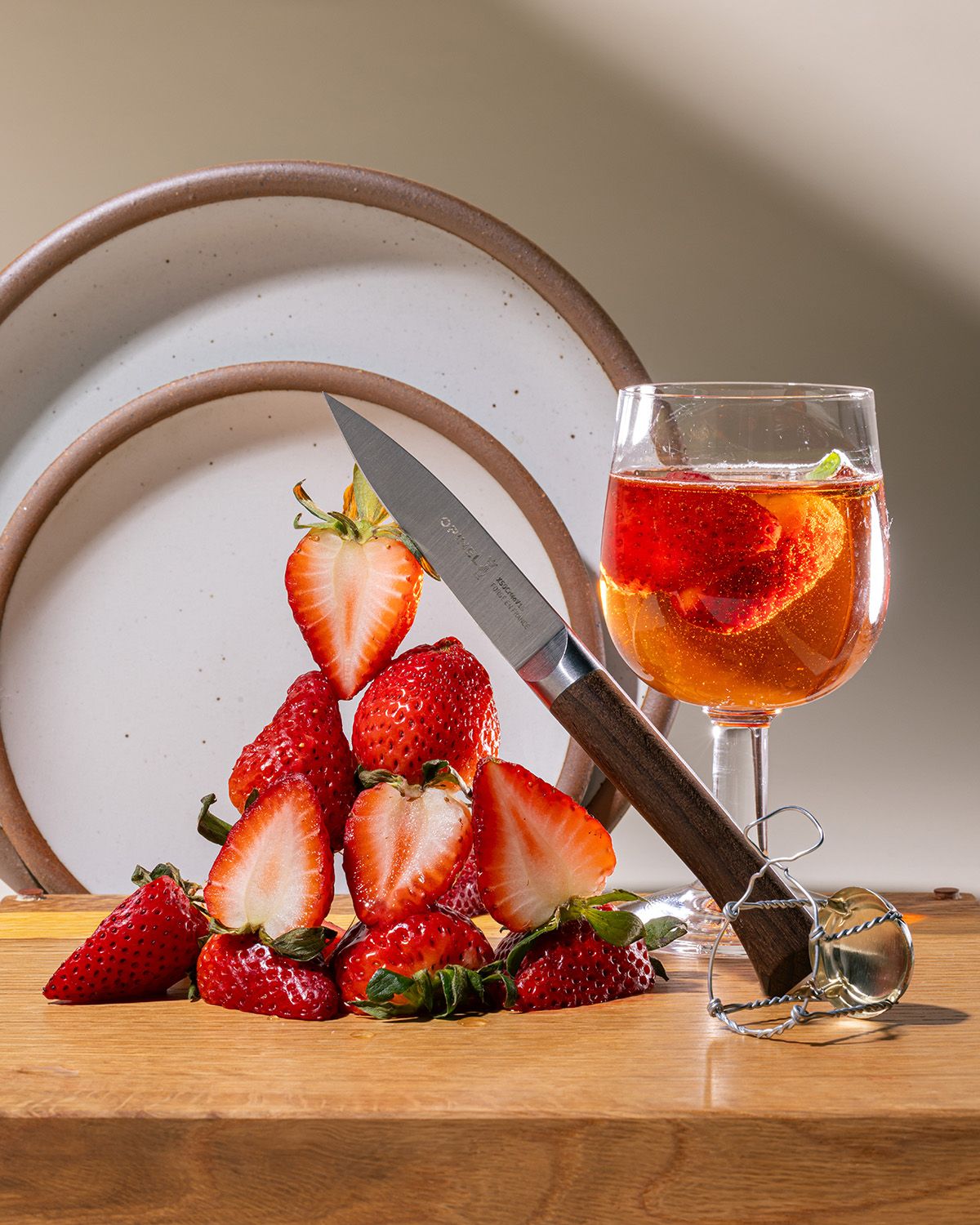 A small paring knife is leaning against a tower of sliced strawberries and sitting on a cutting board. Nearby are a stack of white ceramic plates in the background and a wine glass filled with a sparkling drink and sliced strawberries. The knife has a sharp steel blade and a beveled dark wood handle