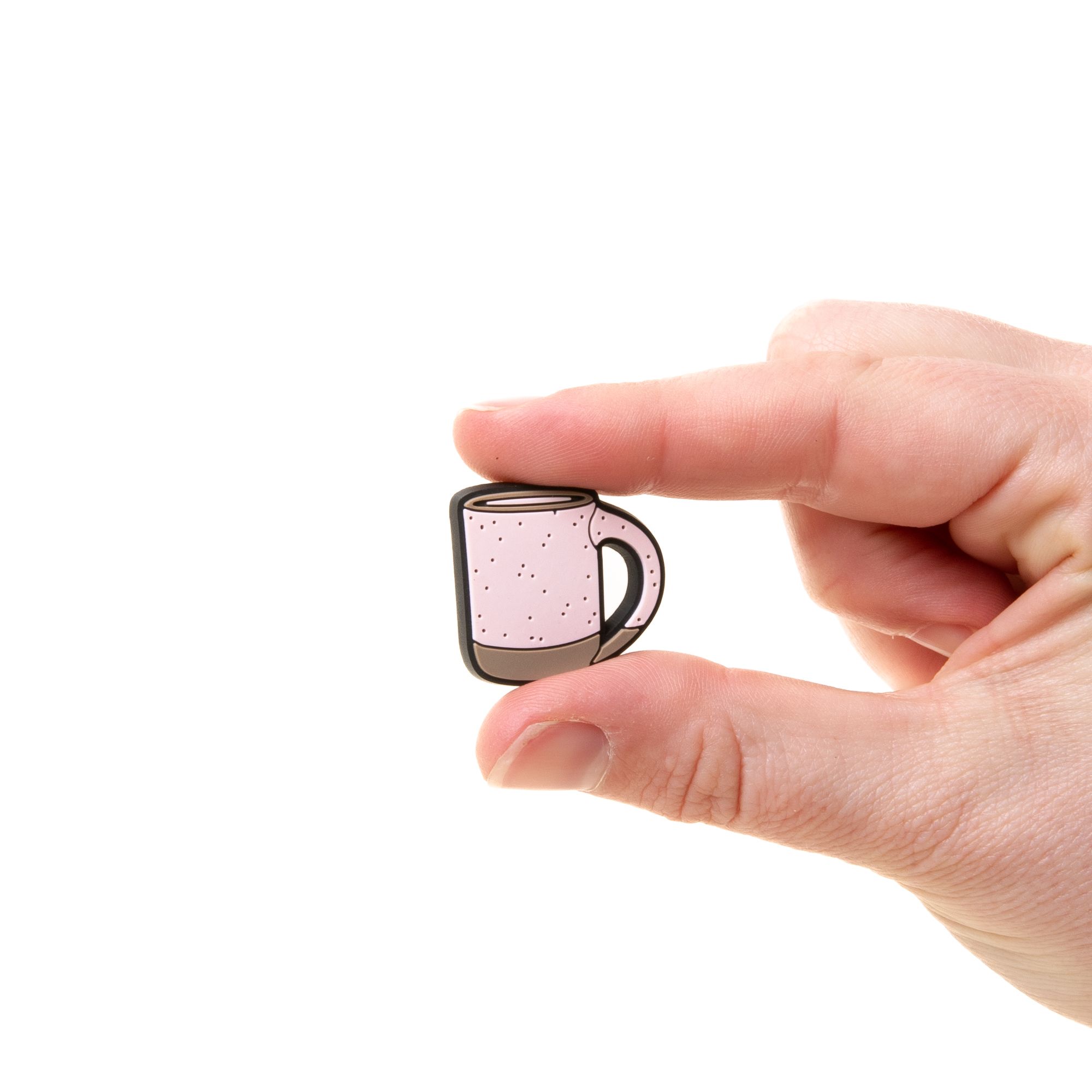 A hand holds a small soft light pink and brown plastic charm shaped like an East Fork Mug for your plastic clogs called Crocs