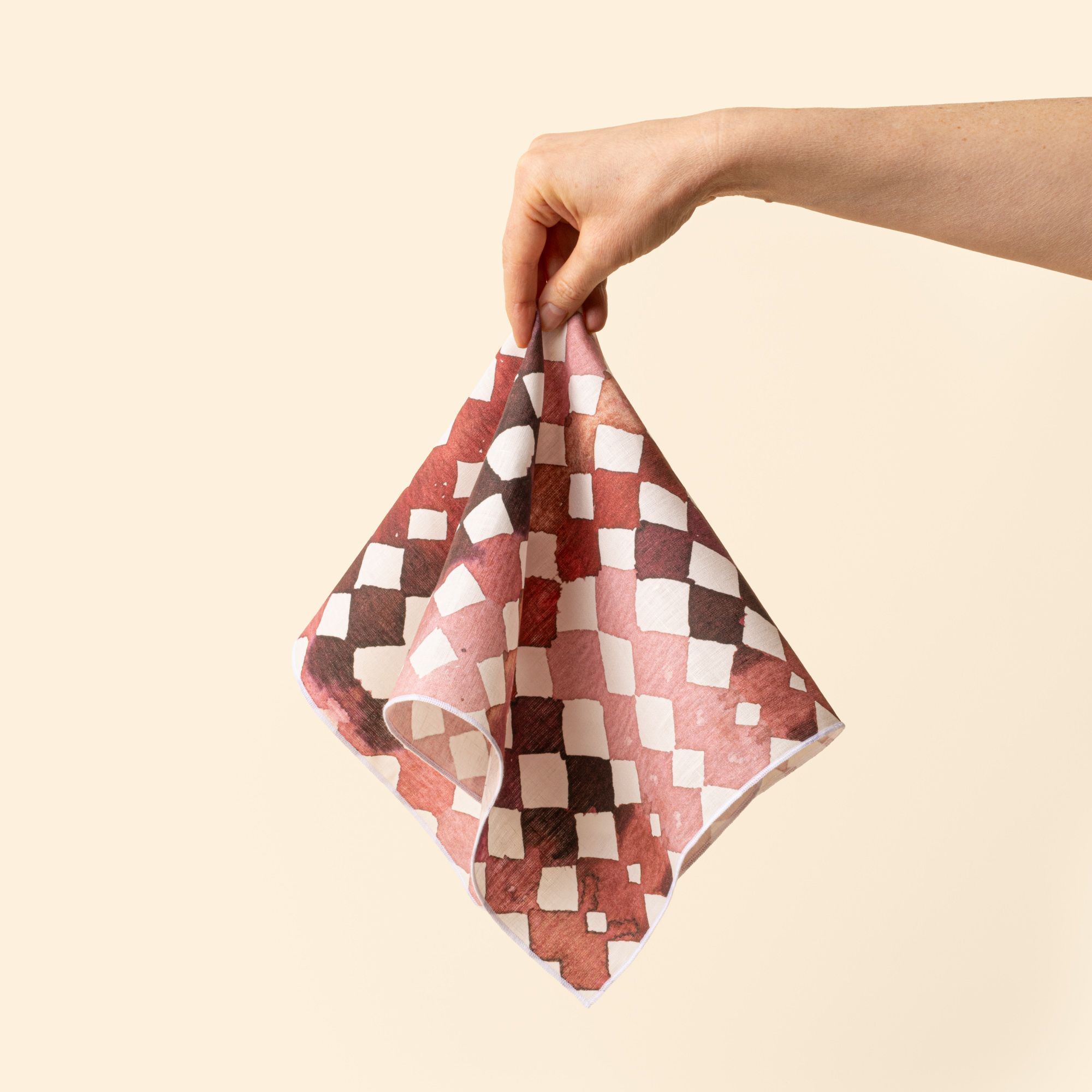A hand holds out a watercolor diamond-patterned cloth napkin in warm burgundy and pink tones
