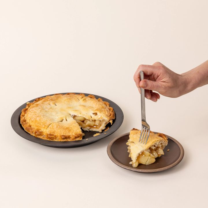 A cooked pie in a simple iron pie plate with a wide rim and shallow body and a slice sitting on a plate next to it. A hand with a fork is about to grab a bite of the slice.