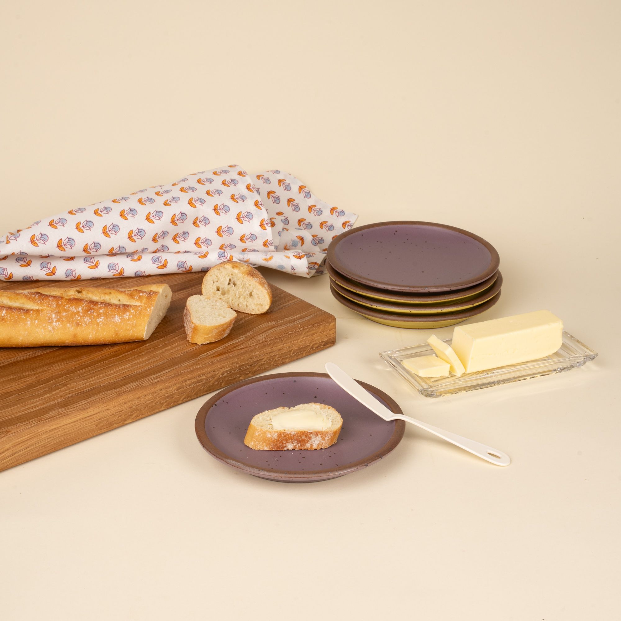 A white enamel butter knife sitting on a plate with a piece of bread and butter, and related items all around.