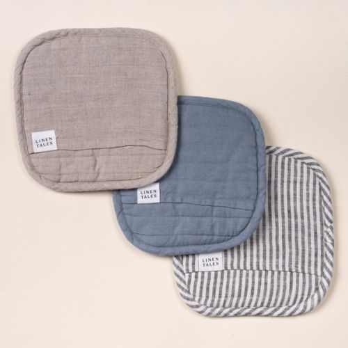 Three square quilted linen potholders in neutral beige, dusty blue, and navy-and-white stripes, each with a small ‘LINEN TALES’ label, arranged overlapping on a light background.