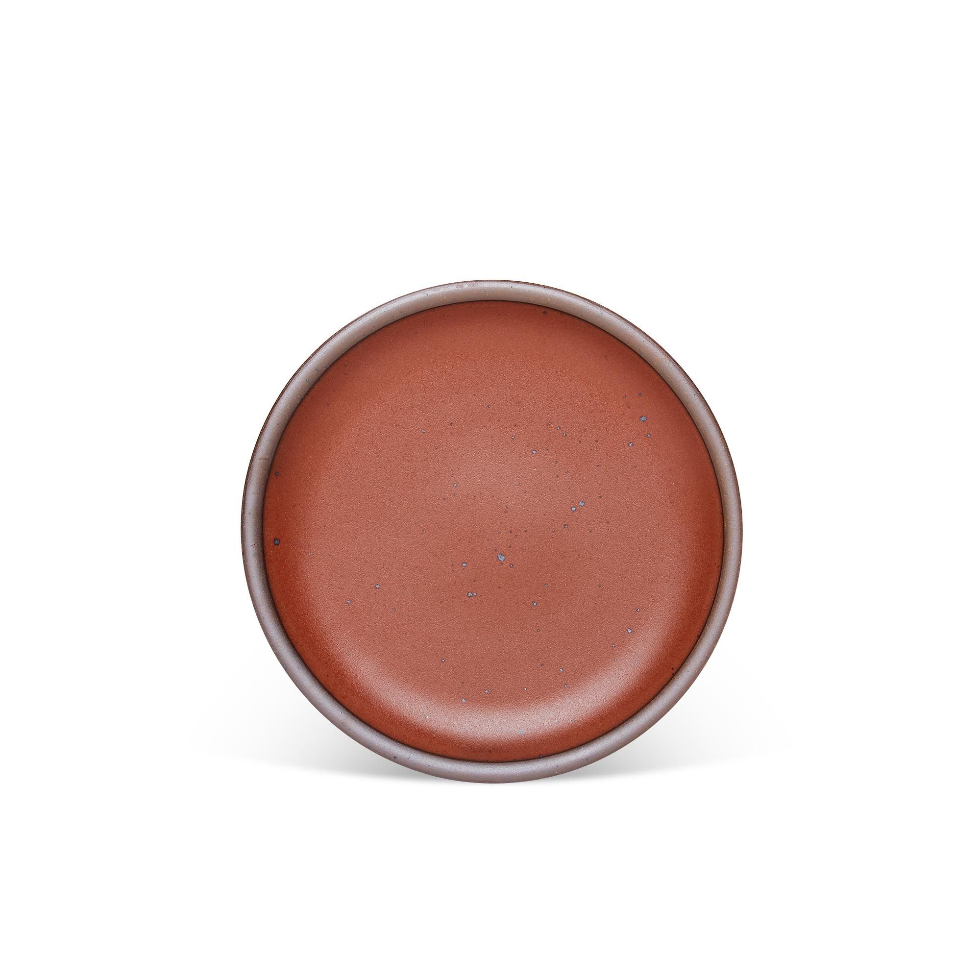 A medium sized ceramic plate in a cool burnt terracotta color featuring iron speckles and an unglazed rim.