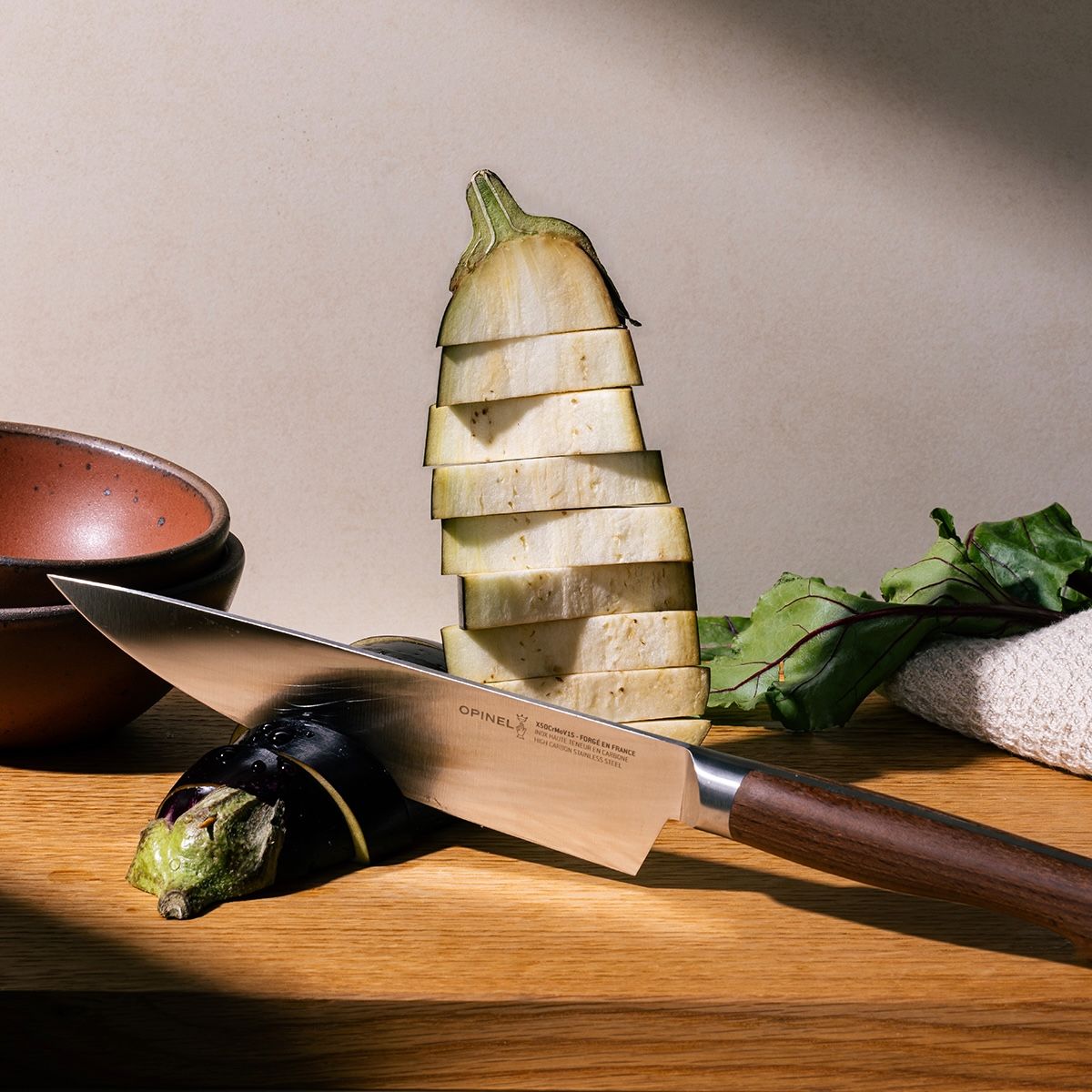 A chef's knife is propped up on a cutting board with slices of eggplant next to the blade and towered behind. The knife has a sharp steel blade with a beveled dark wood handle