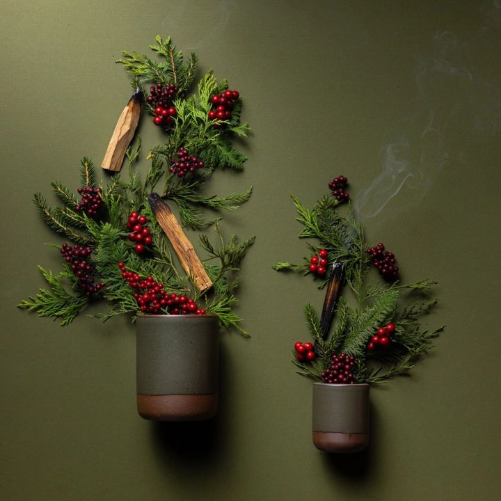 A small and large cylindrical ceramic vessels in a pine green color laying on its side - pine leaves, berries, and cedar are artfully styled coming out of the top of the candles to reflect the scent.