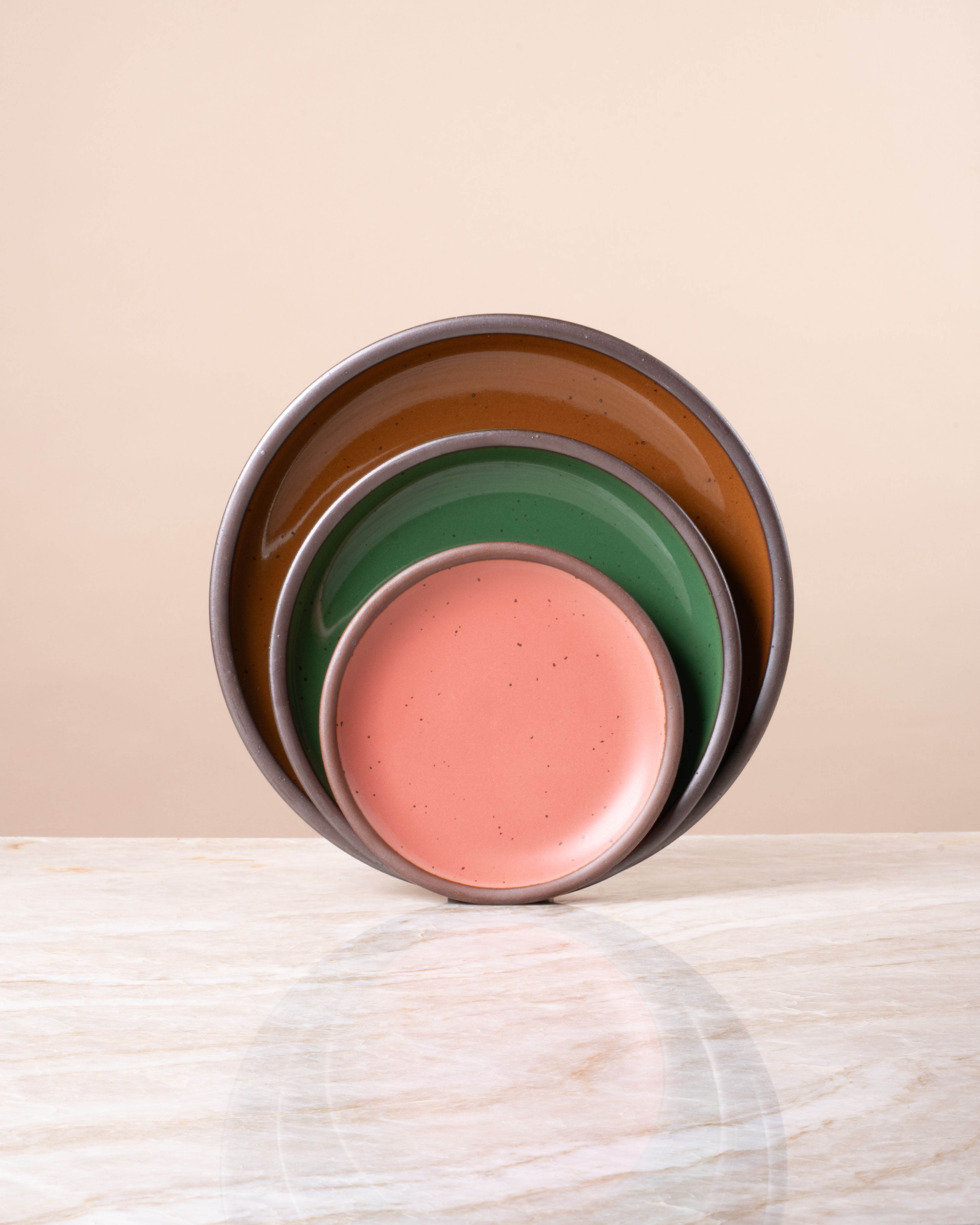 Stacked ceramic plates in glossy green, glossy brown and watermelon pink with speckled finish, displayed on a marble surface.