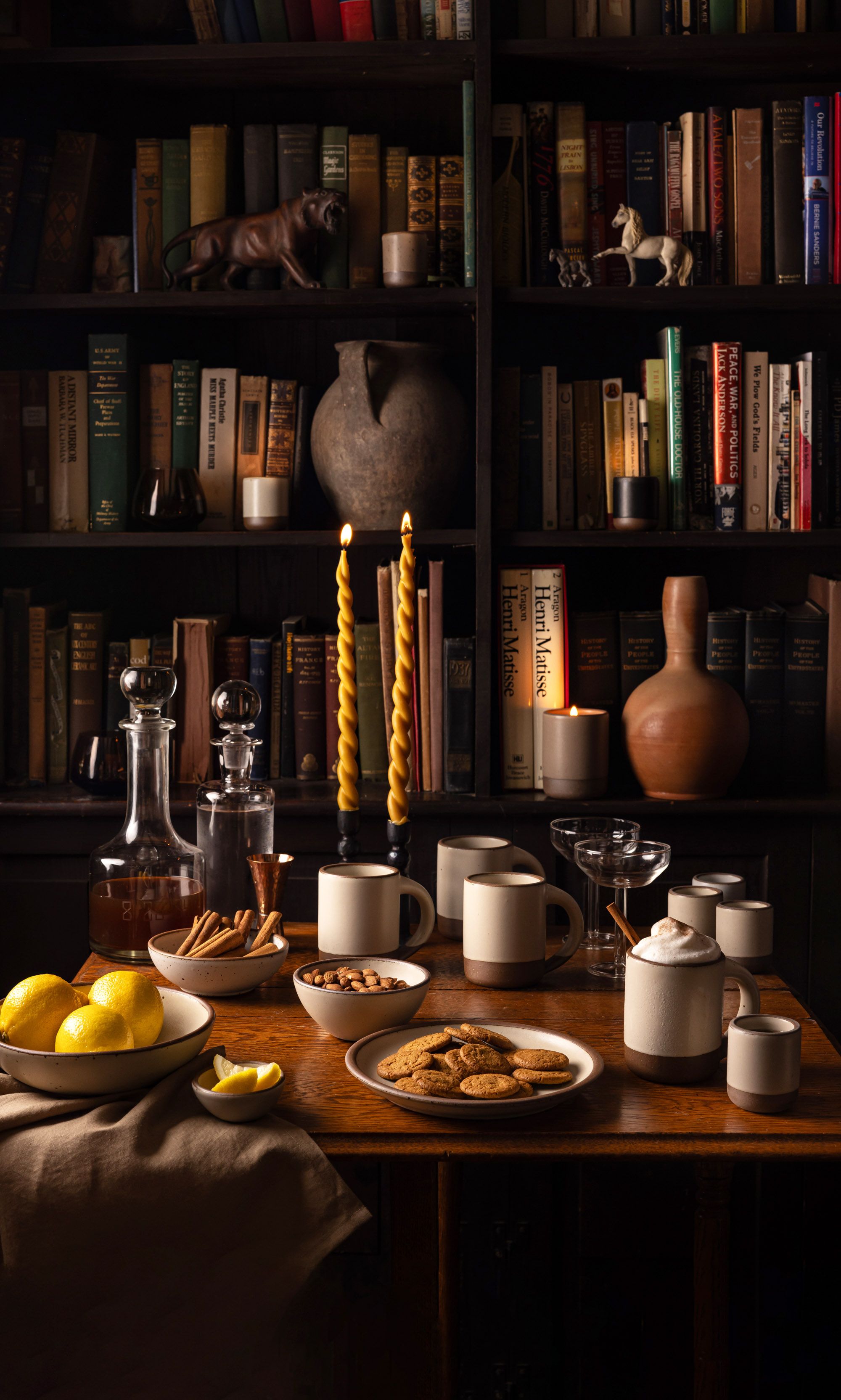 A moody library setting with a table filled with cream ceramic cups, plates, and bowls along with lit candles.