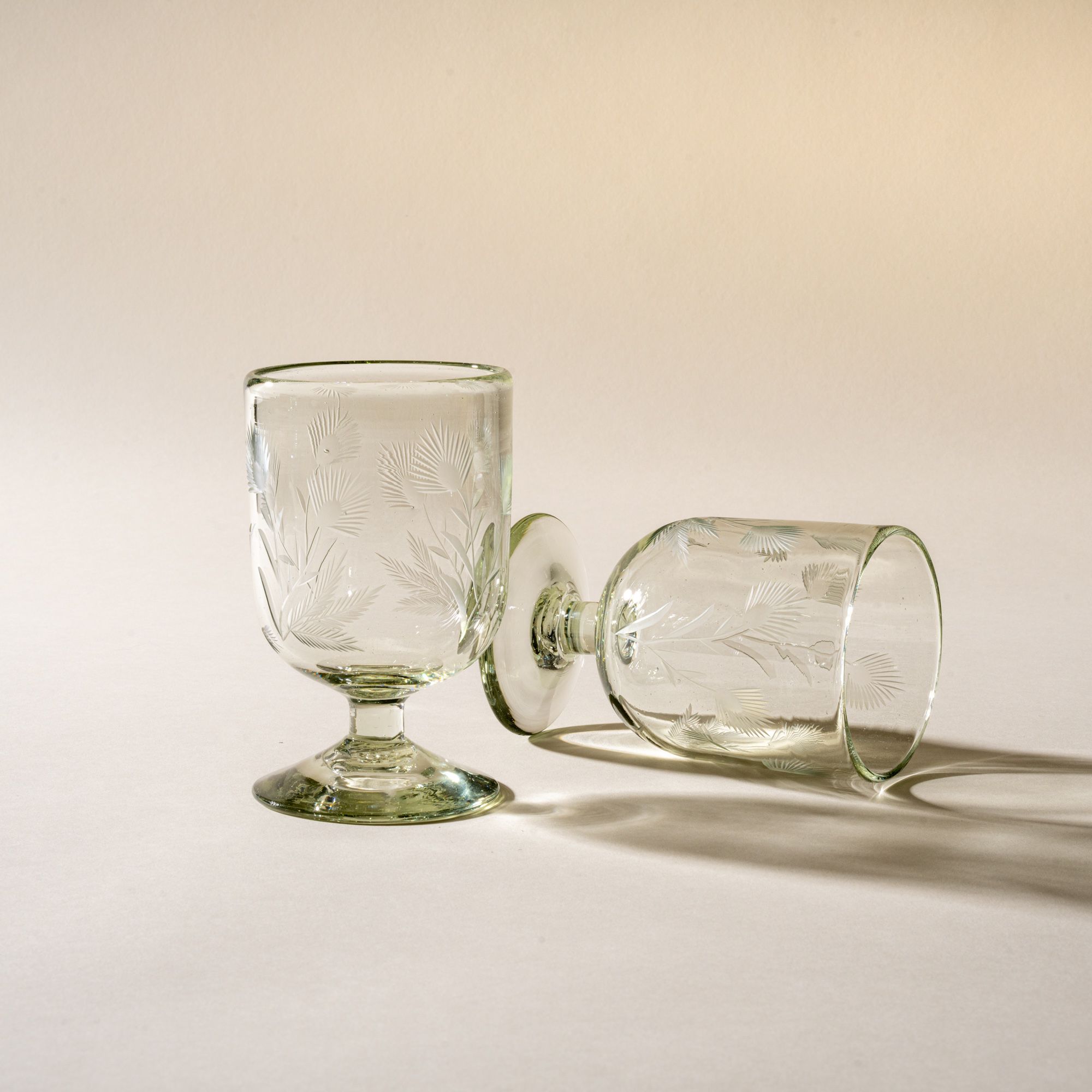 Two clear short wine glasses made of recycled glass with etched thistle designs on it. One is standing up, the other laying on its side.