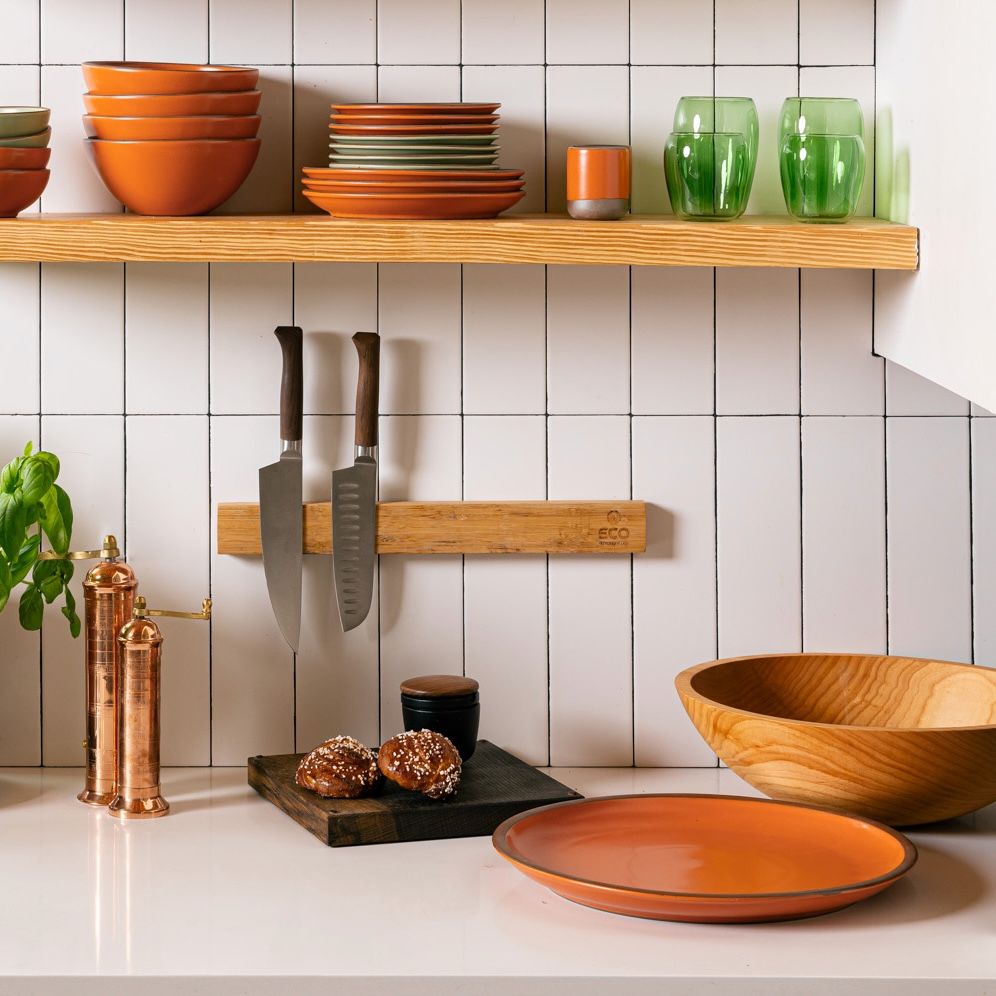 In a kitchen setting, there are floating shelves above a countertop filled with stacks of bowls and plates and green drinking glasses.On the countertop is a large maple bowl, a small dark cutting board, a large circular orange ceramic platter, copper pepper and salt mills, and a knife block on the wall with 2 kitchen knives.