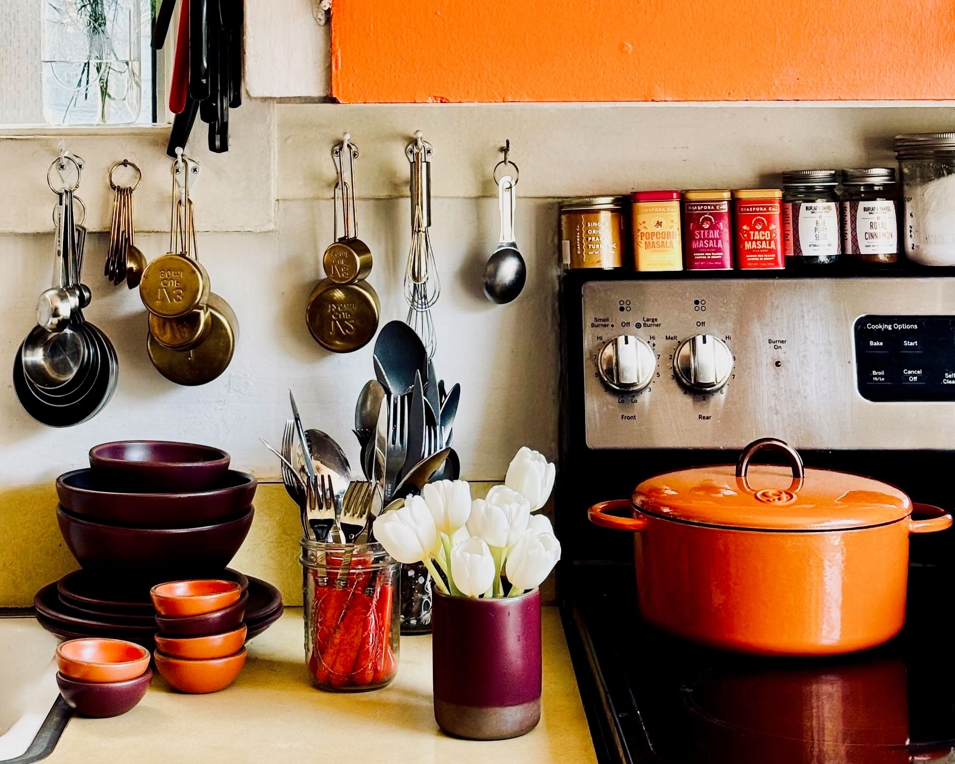 A stovetop and counterspace filled with kitchen tools and colorful ceramic bowls in a bold orange and plum, as well as an orange dutch oven.