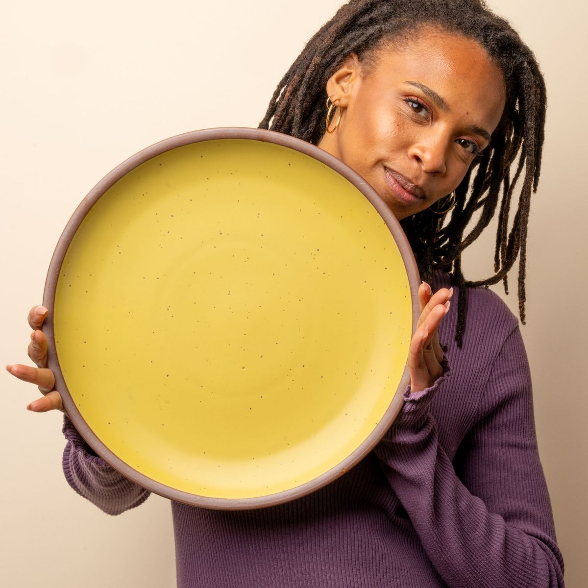 A woman stares at the camera and holds a large ceramic platter in a zesty yellow color featuring iron speckles and an unglazed rim