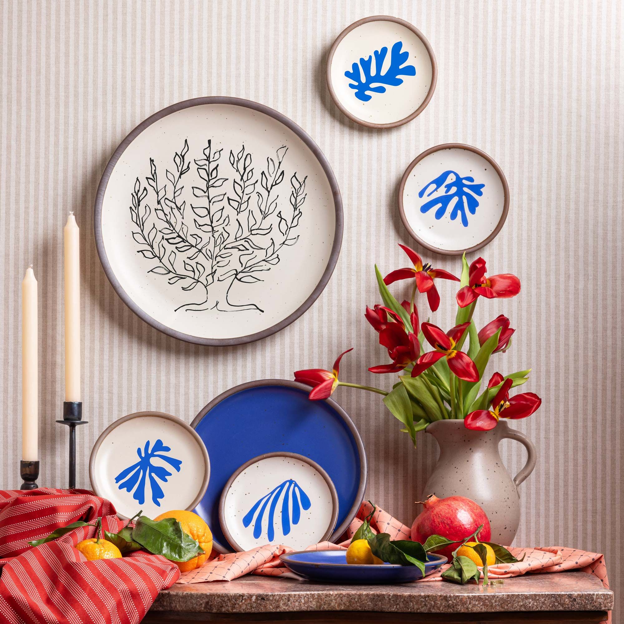 A wall display of several plates featuring Henri Matisse's artworks on the wall along with a cobalt blue plate, flowers, candles, fruit, and red fabric