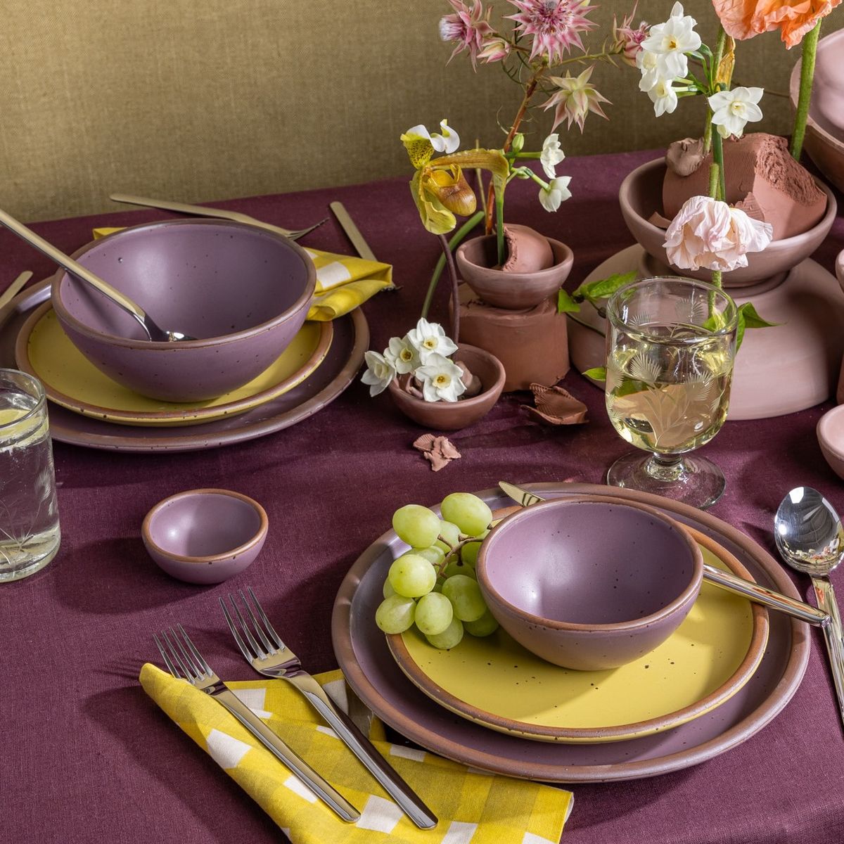 Table settings featuring ceramic dinnerware in soft mauve purple and zesty yellow colors, with silverware, etched glassware, and flowers; all against a purple tablecloth