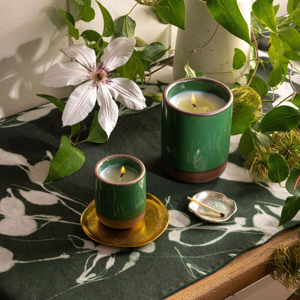 Two glossy emerald green ceramic candles on a floral cloth with a large white bloom, greenery, and a small dish holding a match.
