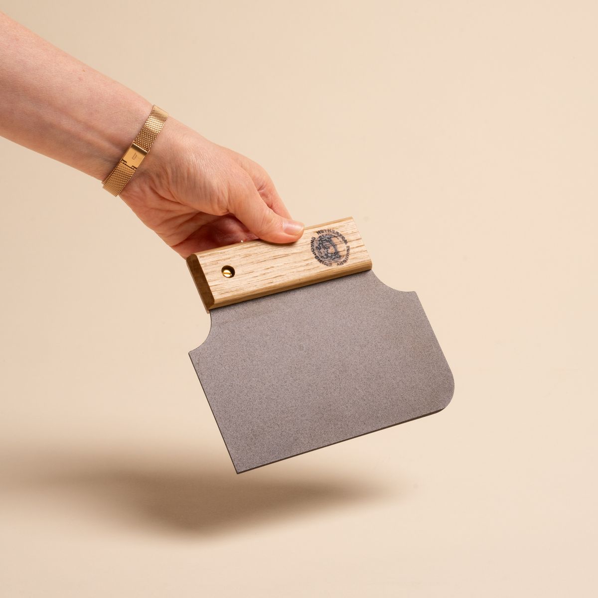 Hand holding a black metal bench scraper with one rounded corner on the blade end while the handle end tapers in to a narrow wooden handle 