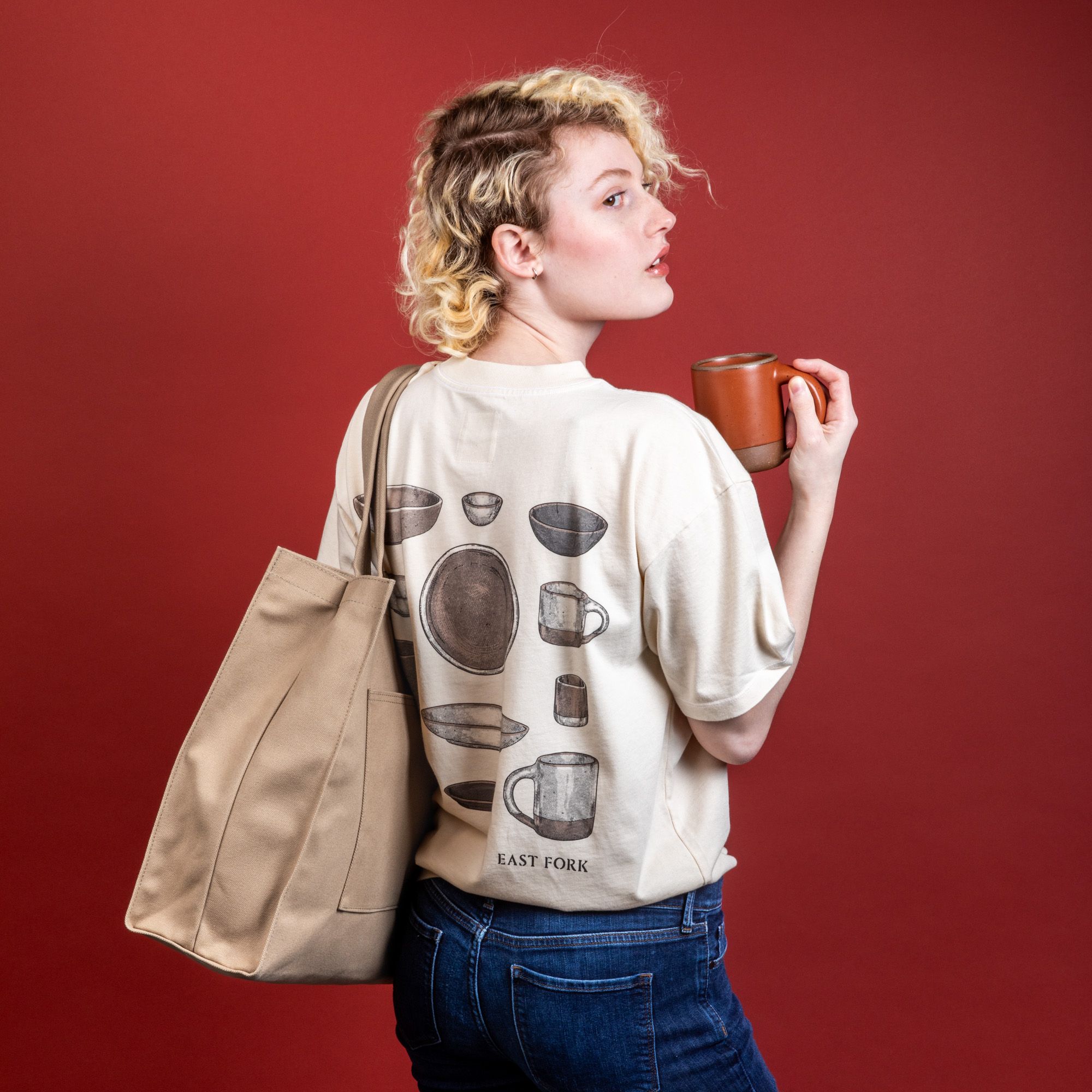 A model wears a t-shirt, jeans, and a sturdy tan canvas tote and a terracotta ceramic mug in her hand.