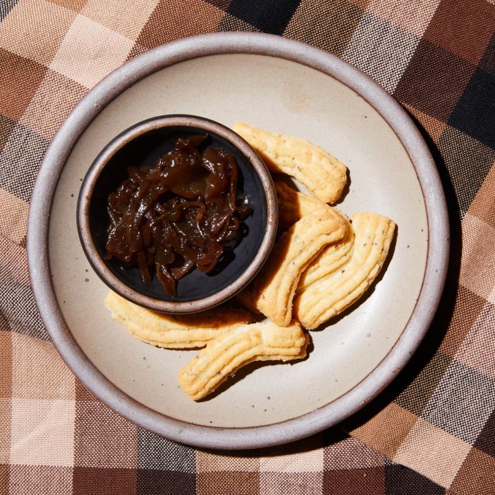 A small bowl of onion jam on a cake plate with crackers on top of a brown gingham tablecloth