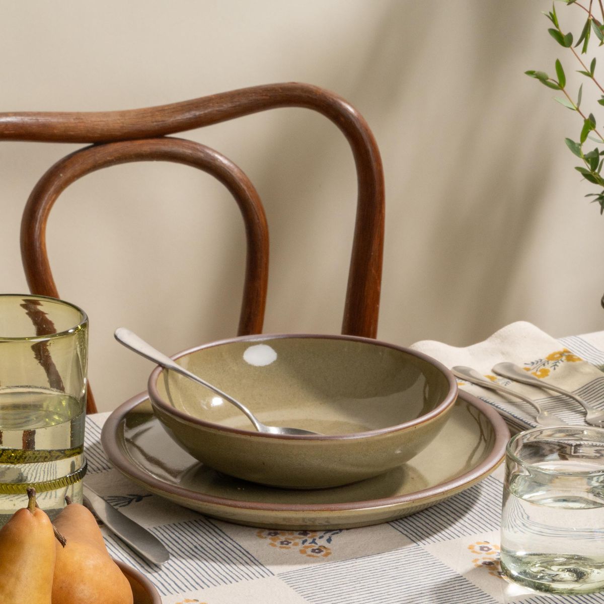 Reflective dappled grey-green stoneware shallow dinner bowl and plate with water glasses and pears on a striped floral tablecloth.