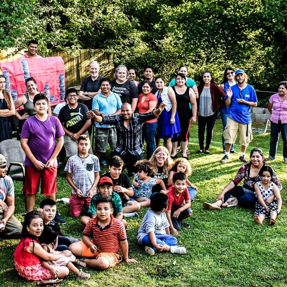 A large group of people sitting and standing outside staring at the camera with green woods behind them.