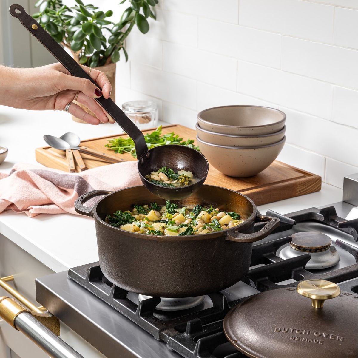 A hand holds a cast iron ladle to scoop out a soup from a cast iron dutch oven on a stove. In the background are soup bowls, spoons, and a kitchen towel.