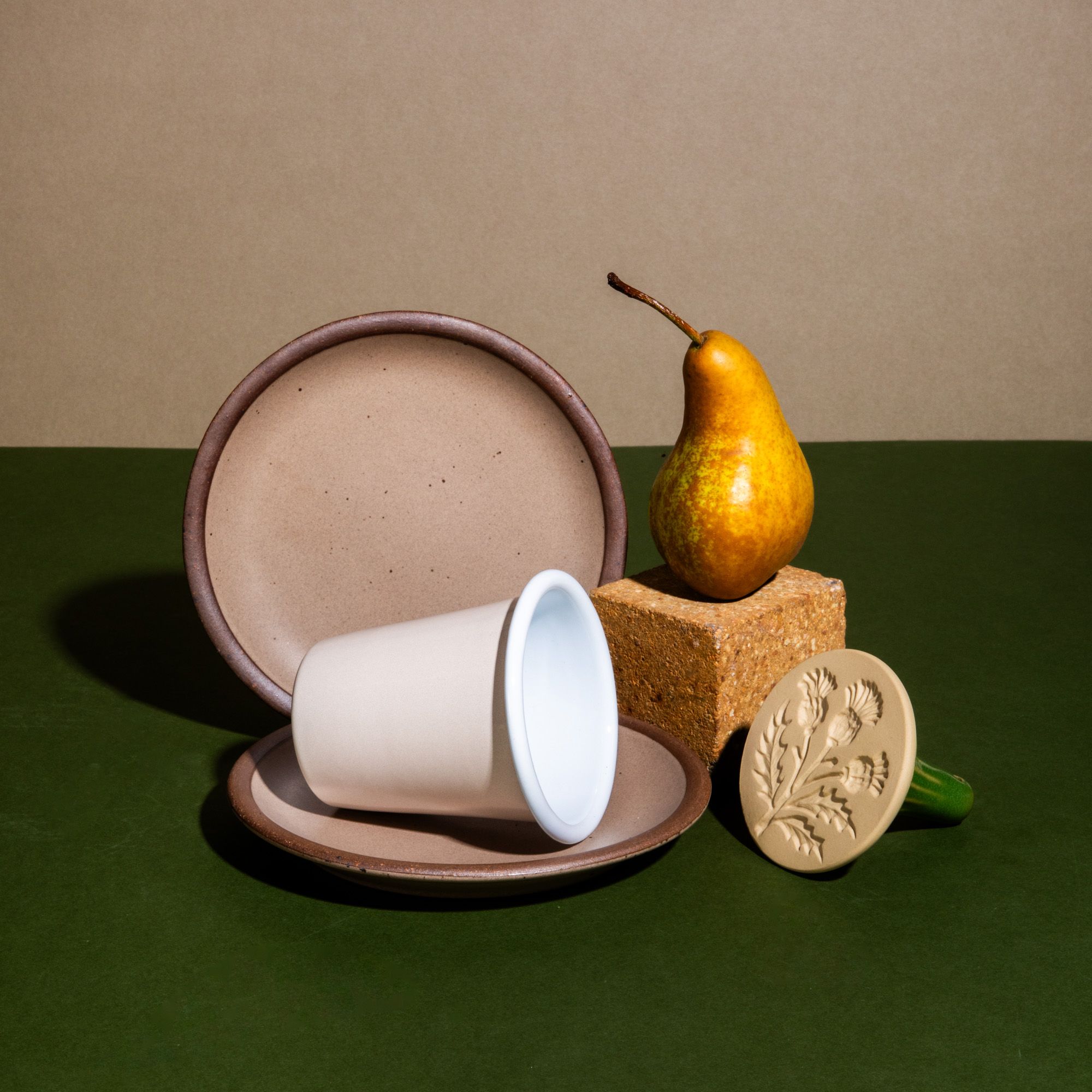 An artful display of light tan ceramic plates, a cream enamel cup, a cookie stamp, and a golden pear arranged against a natural and olive green backdrop.