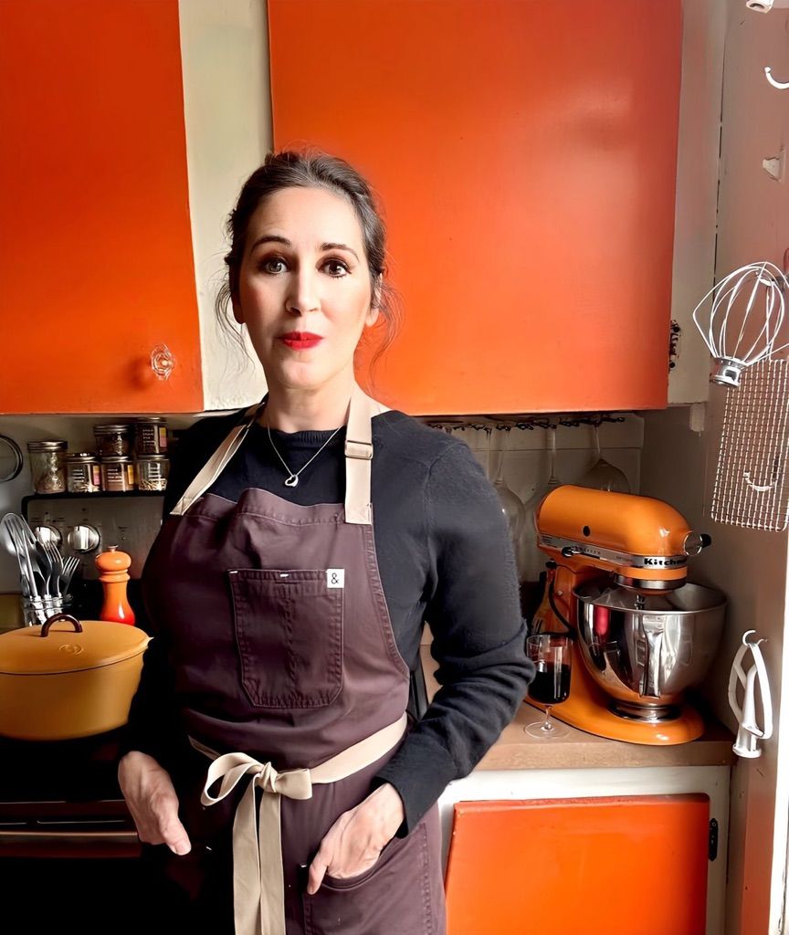 Bebe is wearing an apron and standing in her kitchen featuring lots of orange kitchen tools and orange cabinets