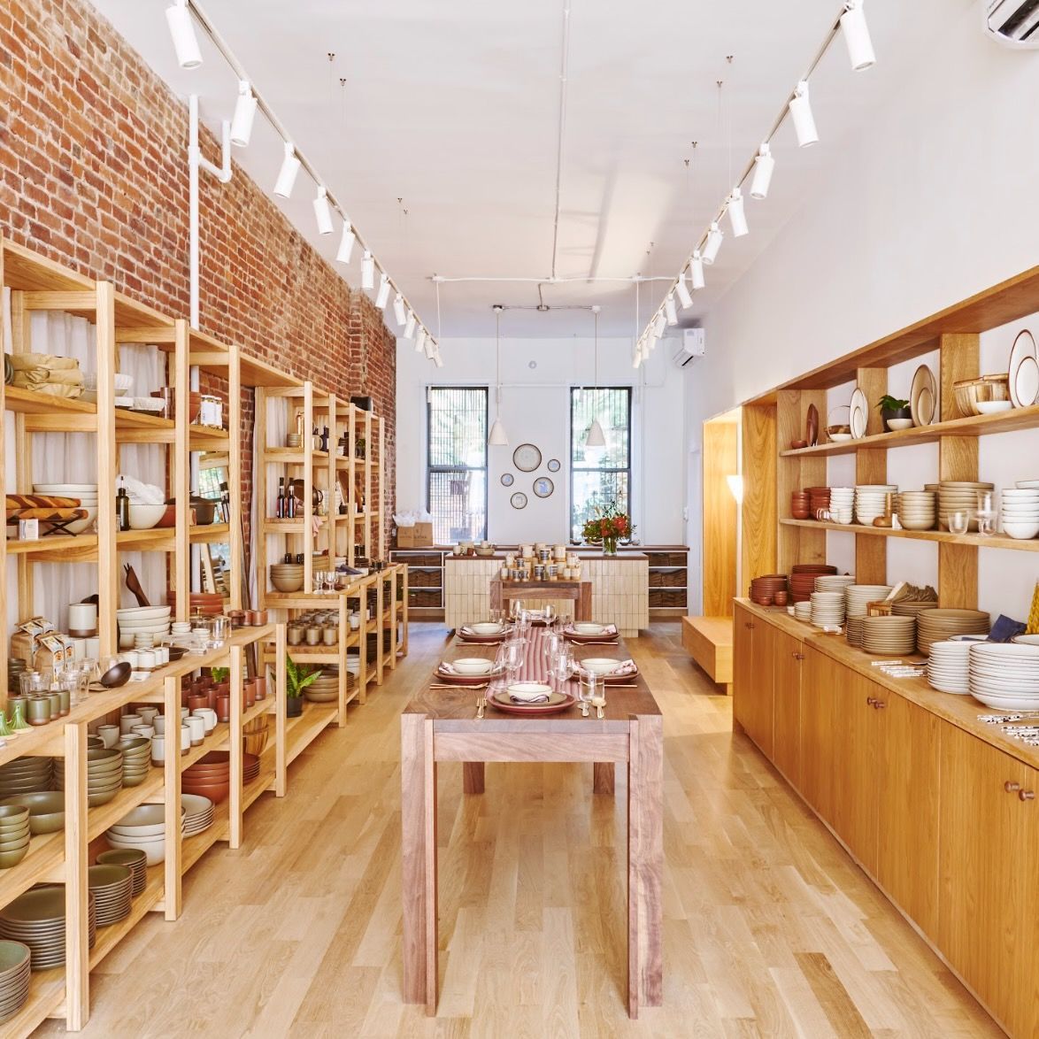 Inside the Brooklyn store location, with a brick wall on the left side and wood shelves lining the left and right walls. A table filled with place setting is in the middle leading to the cash wrap in the back.