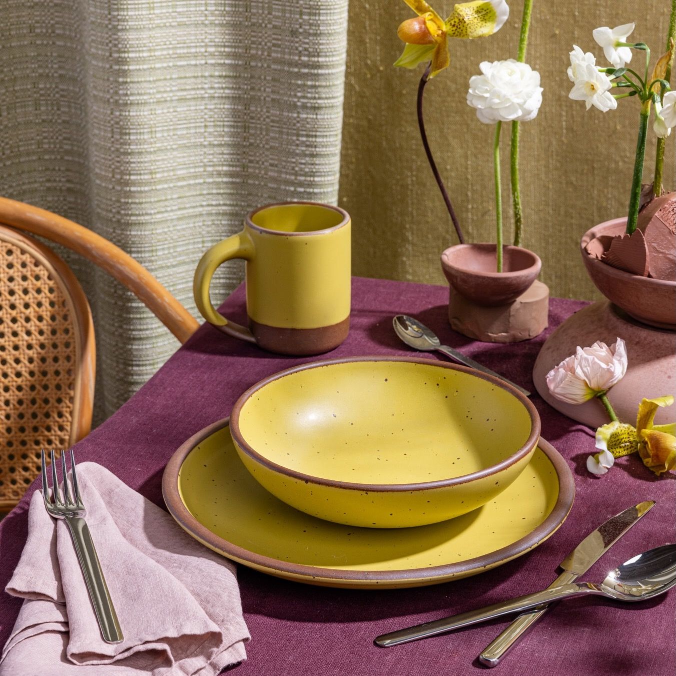 A table setting featuring a zesty yellow ceramic plate, shallow bowl, and mug, along with silverware and a light pink napkin.